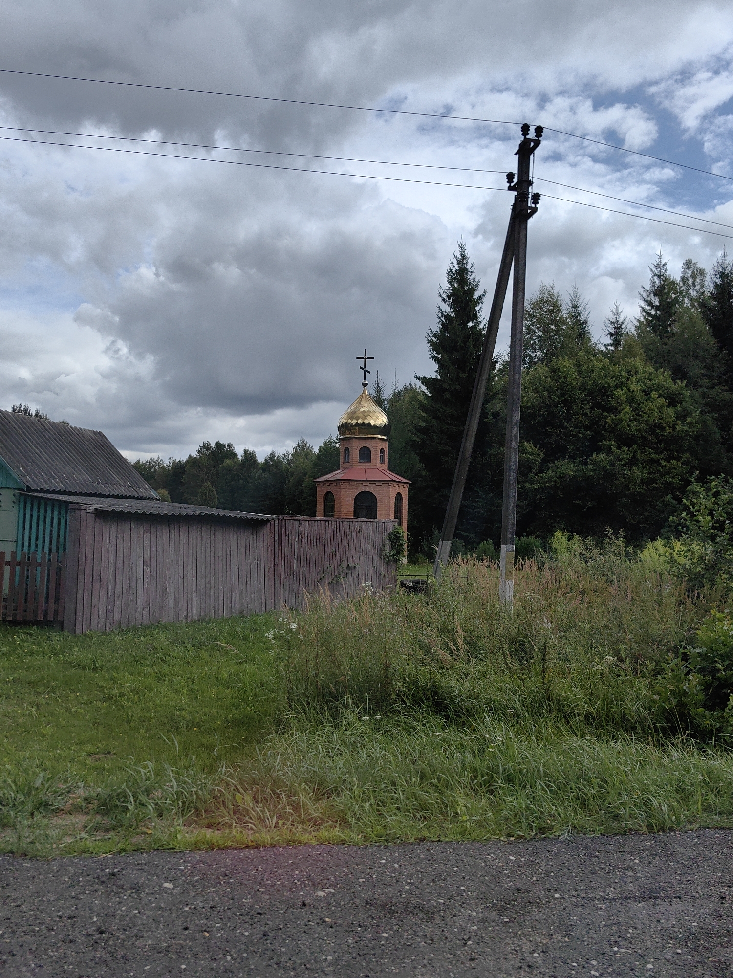 The image depicts an outdoor scene in a rural area, likely a village, with a focus on religious and residential structures amidst natural vegetation. In the background, a small, brick Orthodox church with a golden dome and a cross stands prominently. Adjacent to it, a weathered wooden fence encloses a property. To the left of the fence, a portion of a simple, green-sided building with a corrugated roof is visible. The foreground is dominated by a gravel road, with overgrown grass and weeds lining its edge. A utility pole with electrical wires stretches across the upper portion of the frame. The sky is overcast with heavy, dark clouds, suggesting a possibly stormy or overcast day. The overall impression is one of a modest, possibly remote, rural settlement in Belarus.