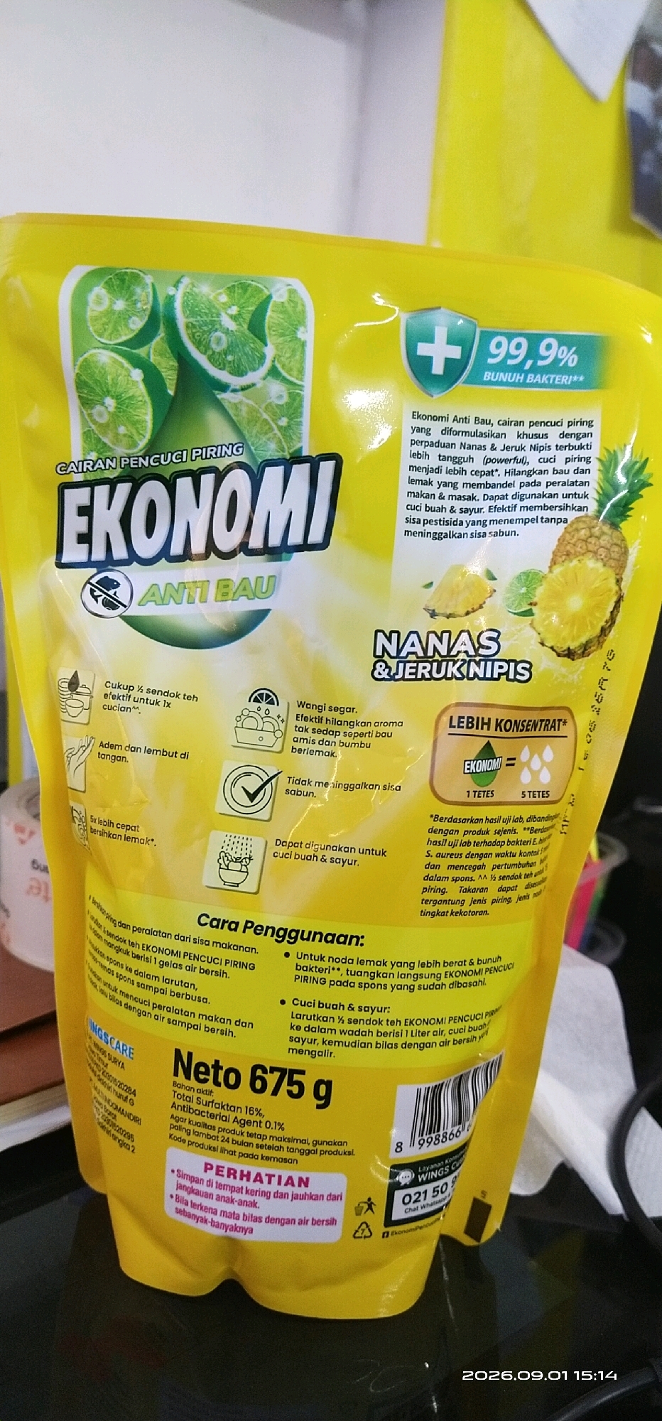 The image shows a close-up of a yellow pouch of "EKONOMI ANTI BAU" dish soap, which is a product of Wings Surya. The pouch features graphics of limes and pineapples, along with text in Indonesian describing its benefits and usage. The product claims to be 99.9% antibacterial and to have a concentrated formula. There is also a "PERHATIAN" (Attention) section warning to keep the product away from children's reach and to rinse eyes thoroughly if contact occurs. The bottom of the pouch displays a barcode and contact information for a customer service hotline. The background is blurred, but suggests an indoor setting, possibly a home or store shelf. No specific location in Tamiang Layang, Indonesia is depicted, as the image focuses solely on the product packaging.