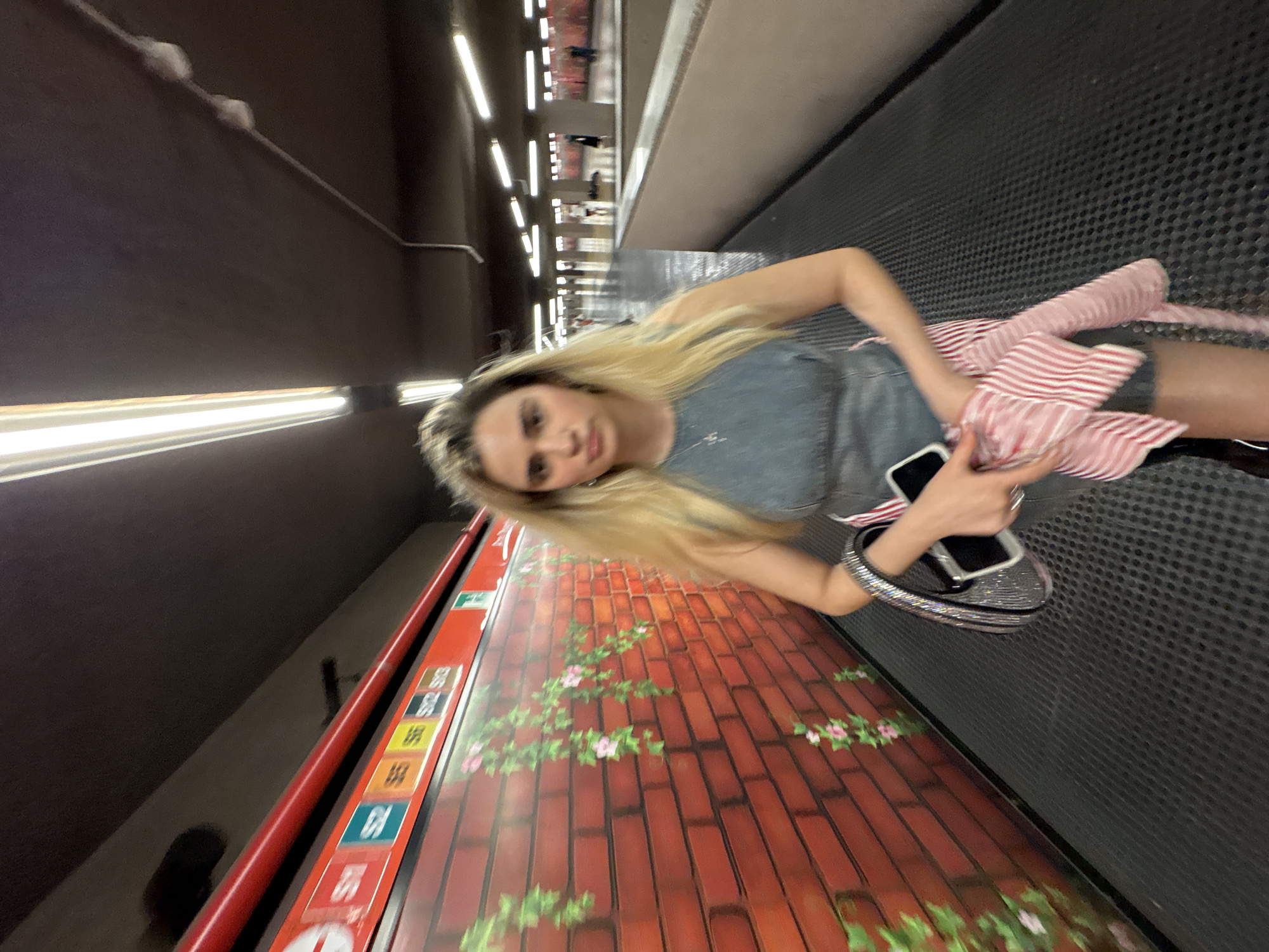 The image shows a blonde woman in Milan, Italy, inside a subway station. She is holding a cell phone in her right hand. She wears a grey shirt, black shorts, and a pink-and-white striped top. The walls and floor of the station are covered with images. The background has an out-of-focus pattern of lights.