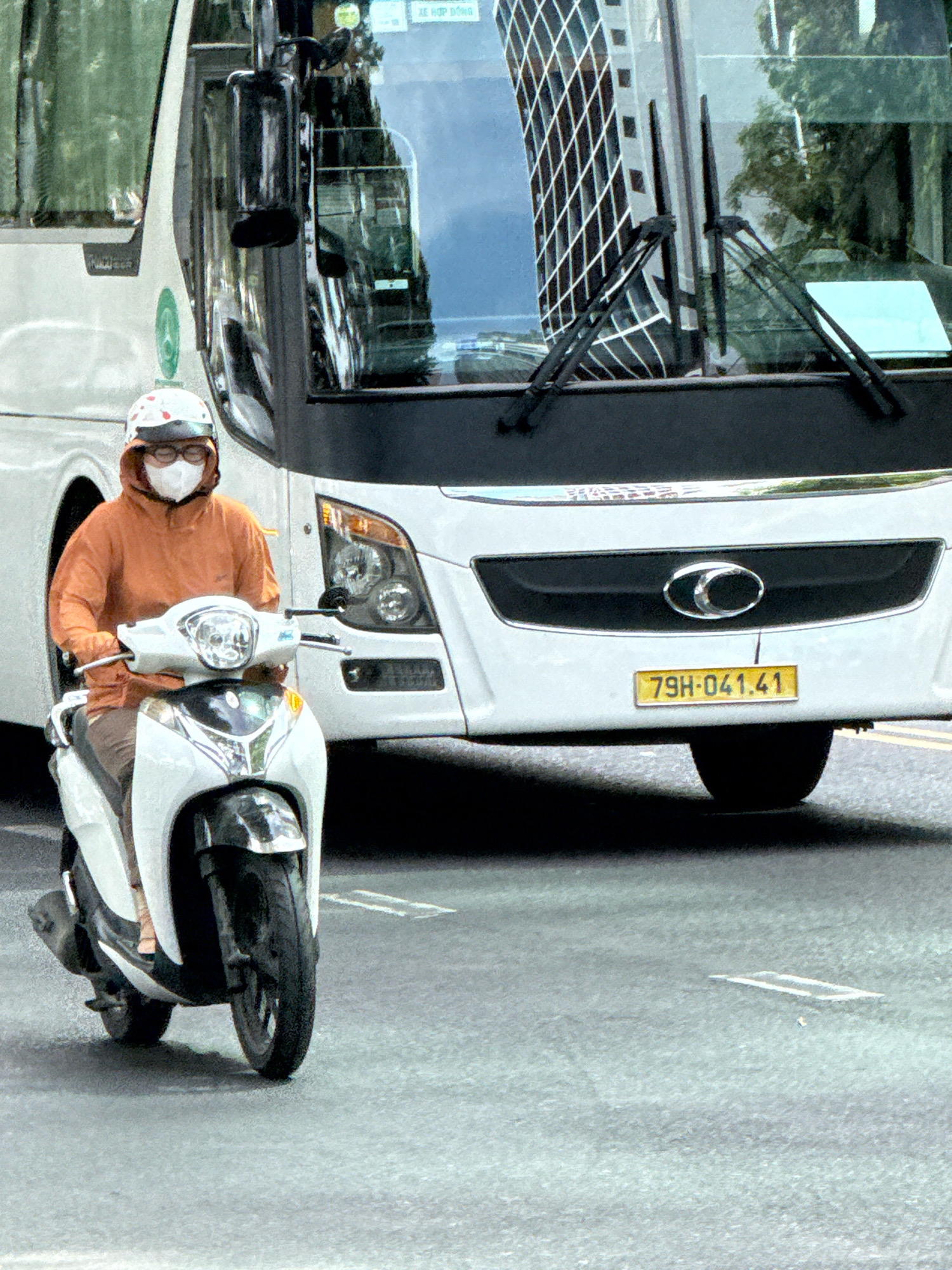 In Nha Trang, Vietnam, a person wearing a white helmet, a face mask, and an orange hooded jacket rides a white and black scooter. They are in the foreground, positioned slightly to the left of the frame. Behind the scooter, a white bus with a prominent black grille and silver trim occupies the middle ground. The bus has its headlights illuminated. A yellow license plate on the bus reads "79H-041.41". The bus's windshield shows reflections of buildings and trees. The road is paved, with white lane markings visible. The environment appears to be an urban setting during daytime, with overcast conditions suggested by the lighting.