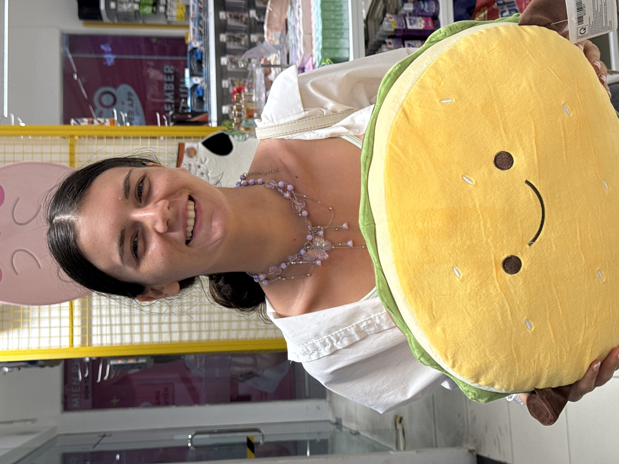 In Phu Thuy Ward, Vietnam, a young woman with dark hair pulled back stands indoors, smiling at the camera. She is wearing a white collared shirt over a light-colored undergarment and a delicate, beaded necklace with a butterfly pendant. She is holding a large, plush taco-shaped toy, which is yellow with green and brown accents, and has a smiling face with dot eyes and a curved mouth. The background shows shelves stocked with various items, suggesting a retail environment. A large, pink cat-ear decoration is visible above the woman's head.