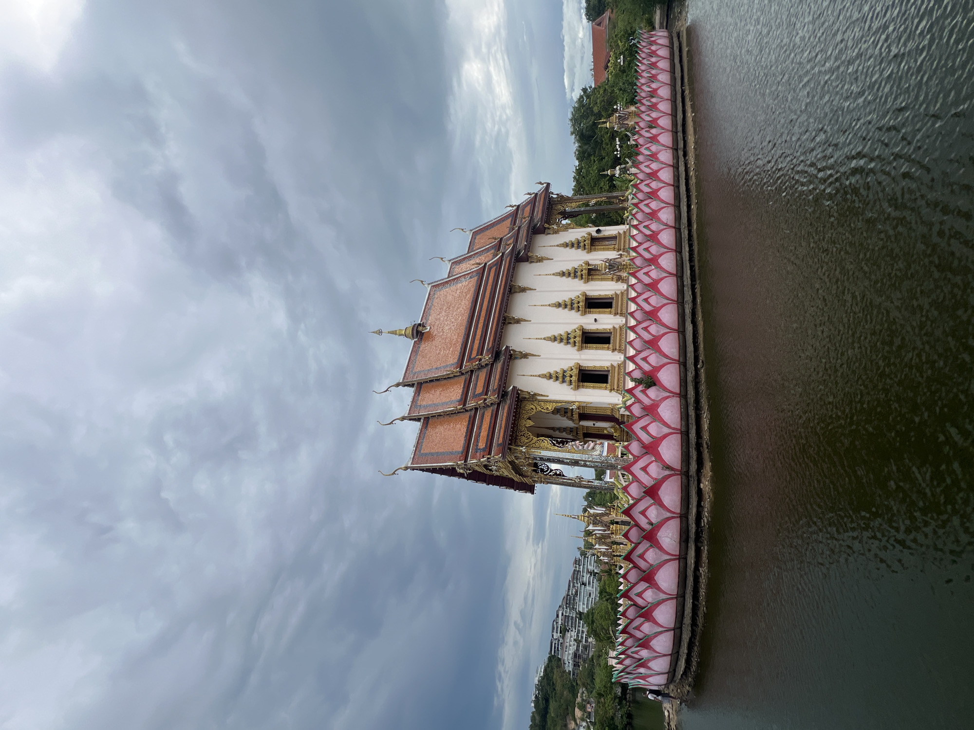 A wide shot captures a Buddhist temple in Ko Samui District, Thailand, situated on a body of dark water. The temple, predominantly white with ornate gold trim and a red tiled roof, is elevated on a platform surrounded by a decorative pink and white lotus-shaped balustrade. The sky above is overcast with heavy, grey clouds. In the background, a lush green hillside is dotted with modern buildings. To the left of the frame, a person stands on the edge of the water, facing away from the camera, wearing a backpack. The water's surface shows ripples and reflections.