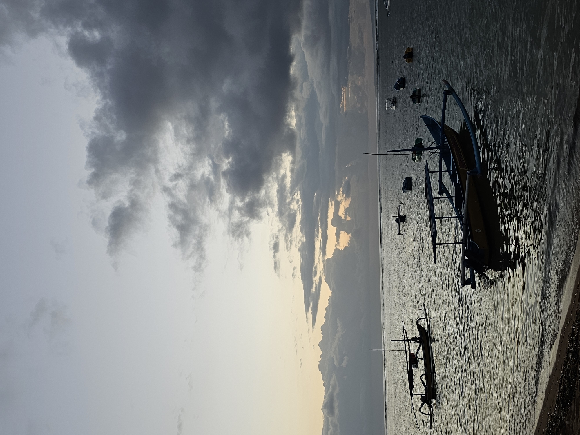 This image captures a serene scene of several traditional outrigger boats (known as jukung in Indonesian) anchored in shallow waters off the coast of Denpasar, Indonesia. The sky is overcast with dramatic, dark clouds, suggesting either dawn or dusk, with a hint of lighter sky peeking through in the distance, possibly indicating the sun is rising or setting. The water is calm, reflecting the muted colors of the sky and the silhouettes of the boats. One prominent jukung in the foreground is yellow and blue, with a canopy and bamboo outriggers. Other jukungs of various sizes and colors are scattered across the water. The shoreline in the lower left corner is visible, consisting of sand and small pebbles. The overall atmosphere is tranquil and suggestive of a quiet morning or evening by the sea.