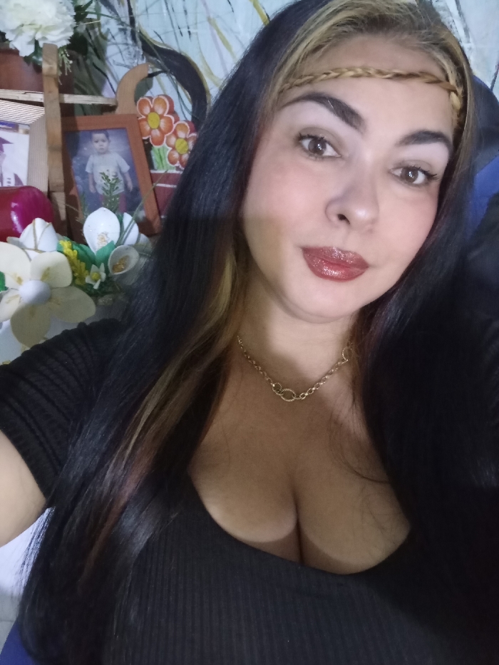 A woman with long dark hair and a braided headband poses for a selfie. She has fair skin, dark eyes, and wears a shiny dark red lip color. A gold necklace adorns her neck. She wears a black low-cut top. Behind her, a small wooden ladder holds a framed photograph of a young child and artificial flowers. Part of a floral patterned wall can be seen.