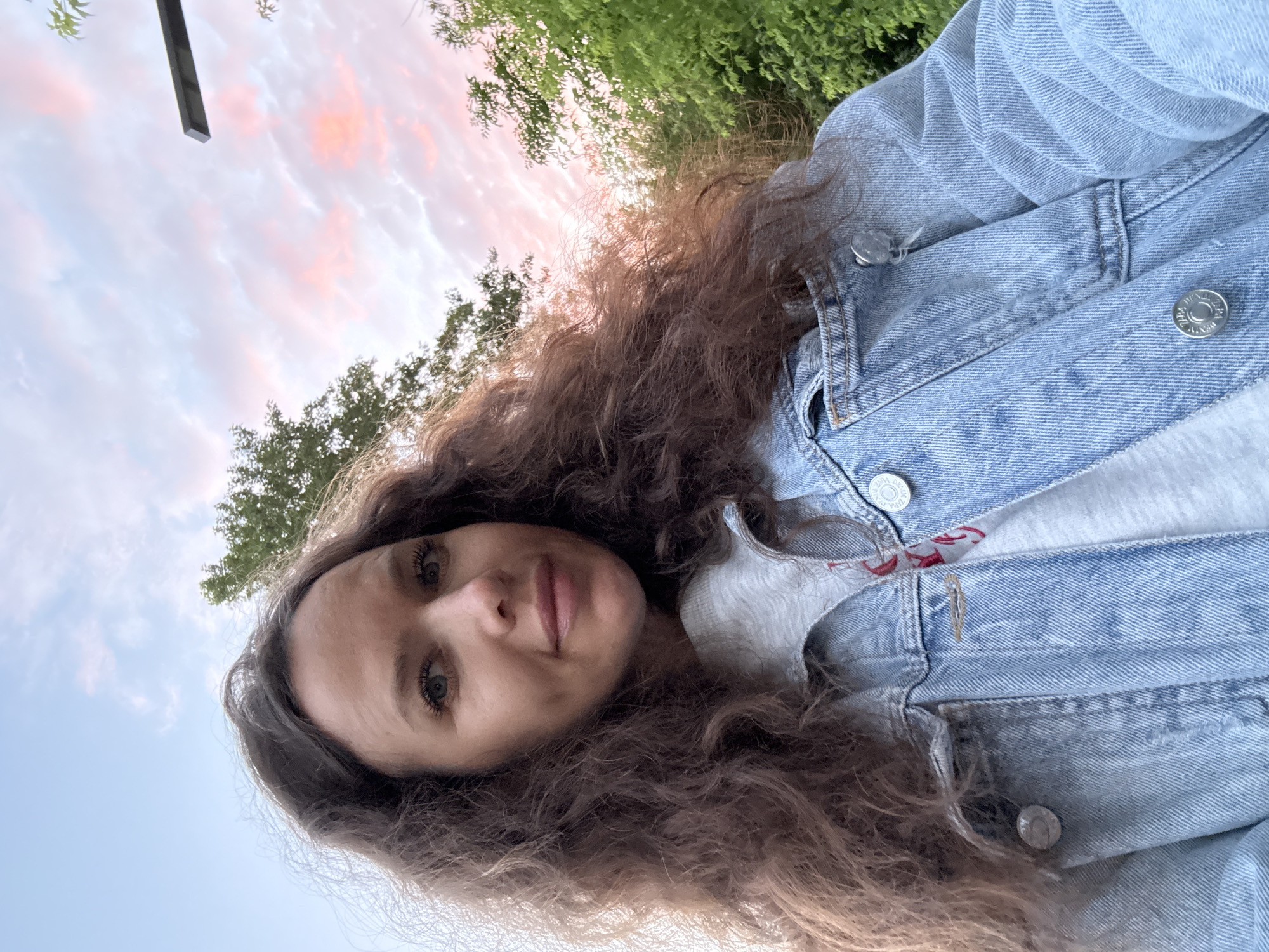 A young woman with long, curly brown hair is taking a selfie outdoors. She is wearing a light blue denim jacket over a grey t-shirt. The background consists of trees and a sky with pink and white clouds, suggesting it is either sunrise or sunset. There is no indication of a specific location within Moscow, Russia.