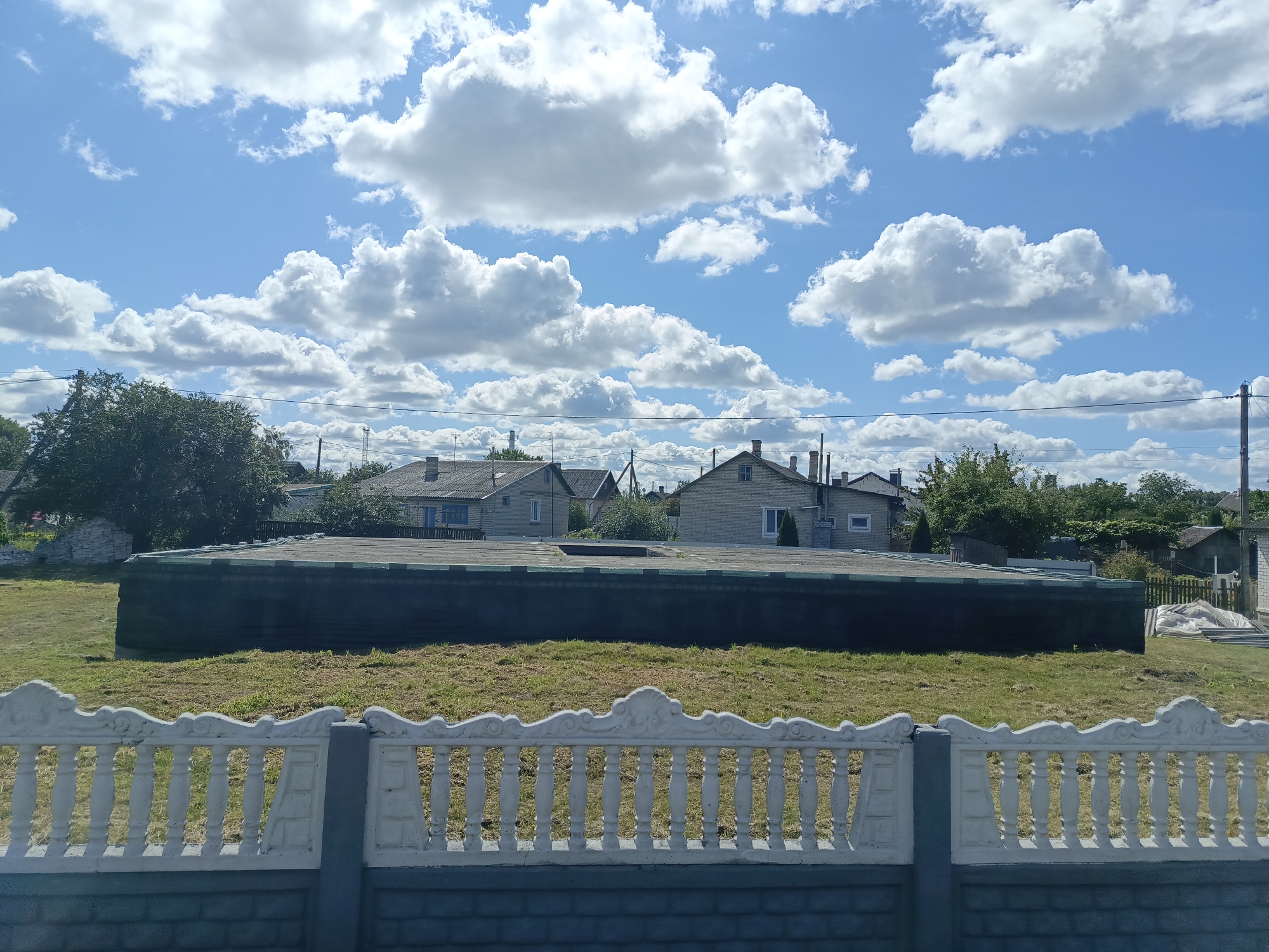 The image depicts a daytime scene in a rural or suburban area, possibly Ivatsevichy, Belarus, characterized by a bright blue sky filled with fluffy white clouds. In the foreground, a decorative white fence with ornate detailing runs horizontally across the frame, resting on a darker grey base. Behind the fence, a flat, dark-colored structure with a visible roofline extends across the mid-ground. This structure appears to be a large, low-lying building or enclosure. Beyond this, a stretch of dry, yellowed grass is visible. In the background, a row of houses with various rooflines and architectural styles are nestled amongst green trees. Power lines are also visible traversing the scene. The overall impression is one of a quiet, sunny day in a residential area.