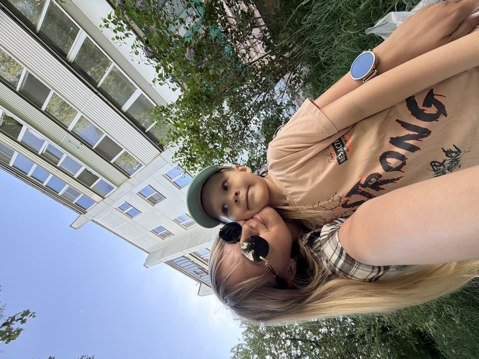 A woman with blonde hair and sunglasses is holding a child with a green cap. The child's face is close to the woman's face. They are outside, with a tall building in the background with multiple windows. There are trees and green foliage around them. The sky is visible and blue. The woman is wearing a plaid shirt and a t-shirt with writing, and she has a watch on her wrist. The location is Chaikovsky, Russia.