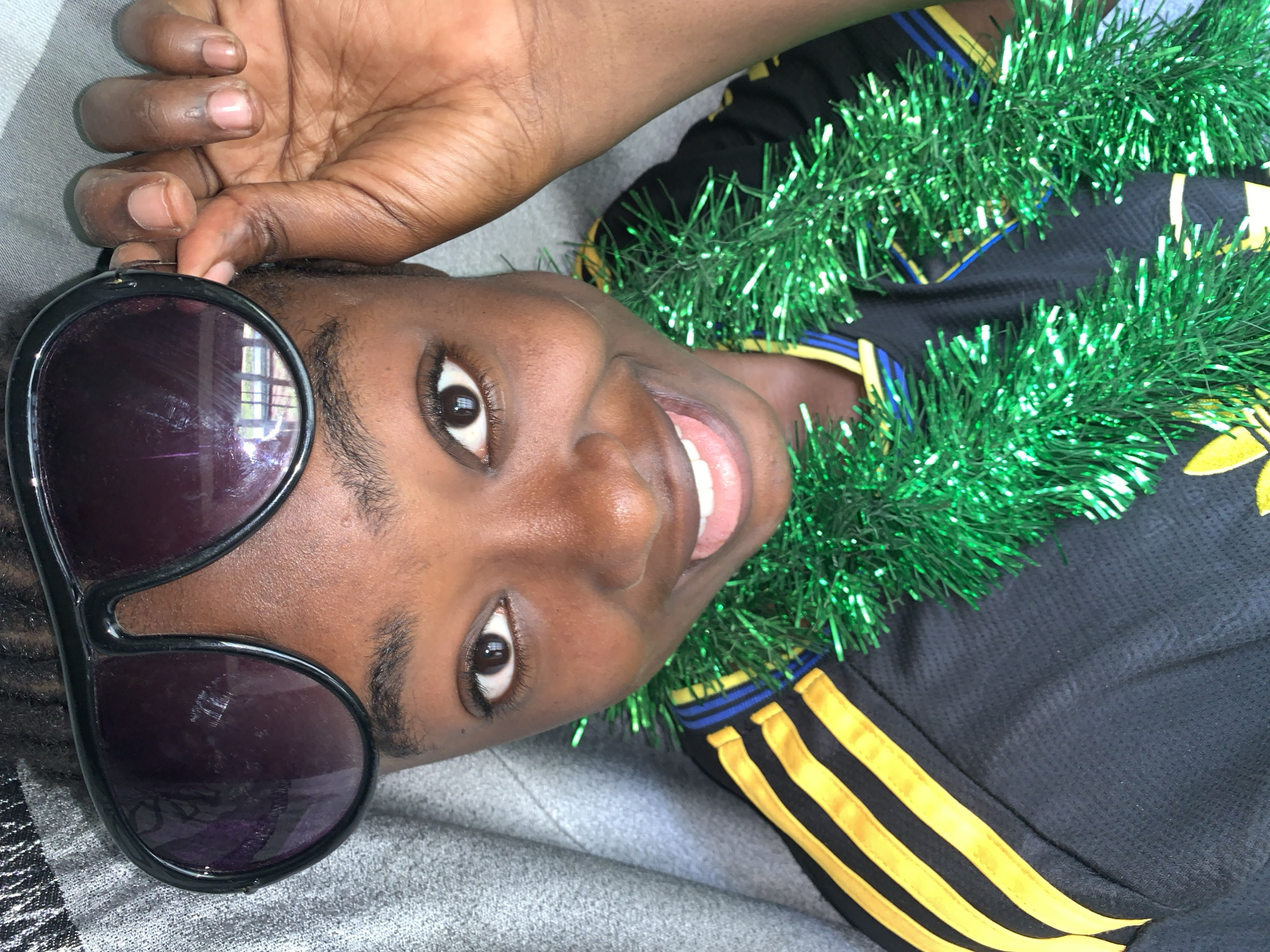 The image shows a close-up, overhead view of a young Black person wearing large, dark-lensed sunglasses and a green tinsel garland. The person is wearing a black athletic shirt with yellow and blue stripes on the sleeve and neckline. Their right hand is raised, holding the side of the sunglasses. They are smiling, with their mouth open and teeth visible. The background appears to be a grey, textured surface, possibly a blanket or upholstery. The lighting is bright, casting subtle shadows. There are no discernible elements indicating a specific location within Rafin Par, Nigeria.