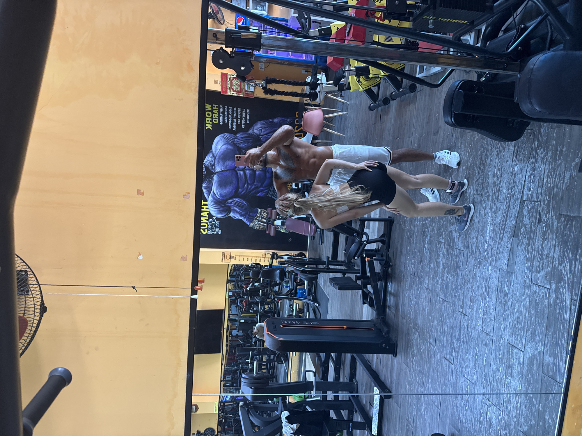 In a gym located in Nha Trang Ward, Vietnam, a man and a woman are taking a mirrored selfie. The man, shirtless and with defined abs, wears white shorts and is holding a phone to take the picture. The woman, with long blonde hair, wears a white sports bra and black shorts. They are positioned in front of a wall with a large mural of a muscular blue figure. The gym appears to be well-equipped with various weight machines and exercise equipment visible in the reflection and around them. The floor is made of grey wooden planks, and the walls are painted a light yellow.