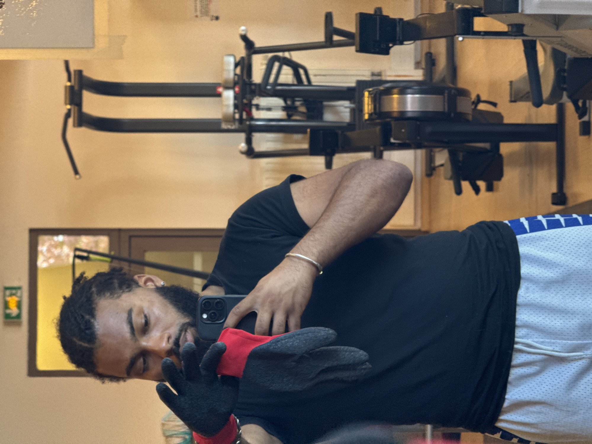 A man with braided hair and a beard, wearing a black T-shirt and blue and white athletic shorts, is taking a selfie in front of a mirror. He is wearing a red and black glove on one hand. In the background, a weight training machine is visible. The photograph appears to be taken indoors, likely in a gym or fitness room. The location is unknown, but it is unlikely to be in Sarcelles, France because there are no indications of this.