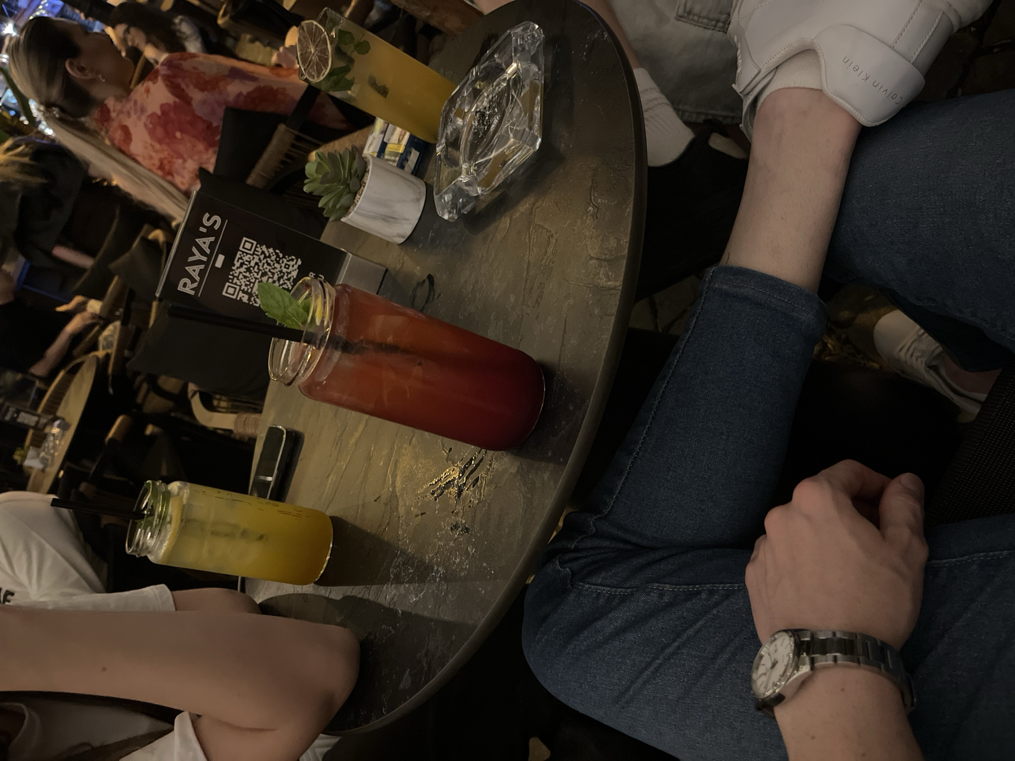 The image captures a high-angle view of a table at RAYA'S bar in Plovdiv, Bulgaria. Two people are seated at the table. One person wears blue jeans and white Calvin Klein sneakers. Another person wears a white top. On the table are two drinks in mason jars, one yellow and one red, an ashtray, a small potted succulent, and a RAYA'S branded menu or display with a QR code. A person wearing a dress with floral print is also visible in the background. Another person with a wristwatch on their left wrist sits at the table. The table is dark and textured. The overall lighting is dim.