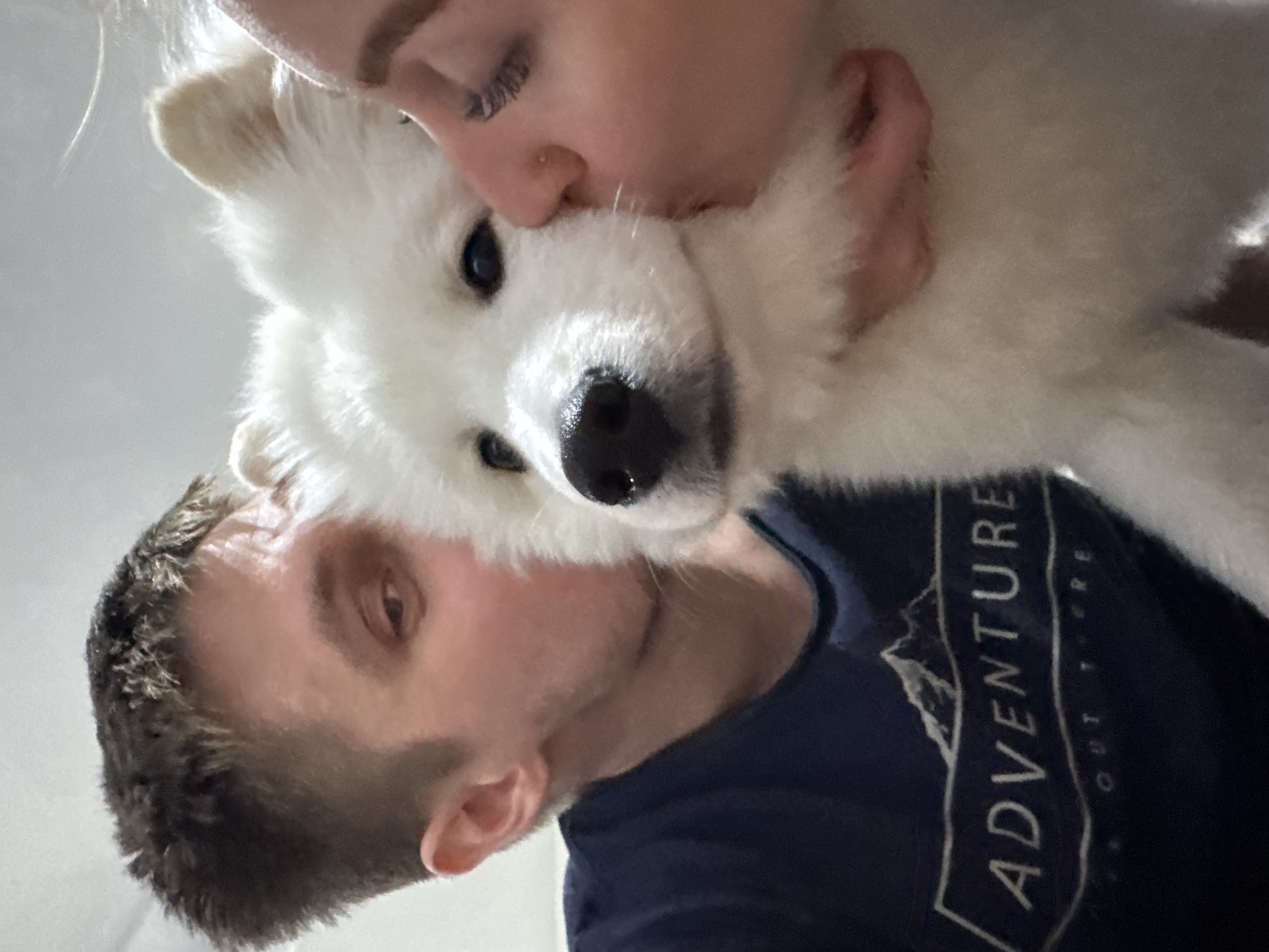 A close-up, angled selfie shows a white Samoyed dog being held by two people. A woman with blonde hair and a nose ring kisses the dog's head. A man with short brown hair and a blue shirt with the word "ADVENTURE" is visible below the dog. The background is a plain white wall. The scene appears indoors. There is no information regarding the location in the city of Chaikovsky, Russia.
