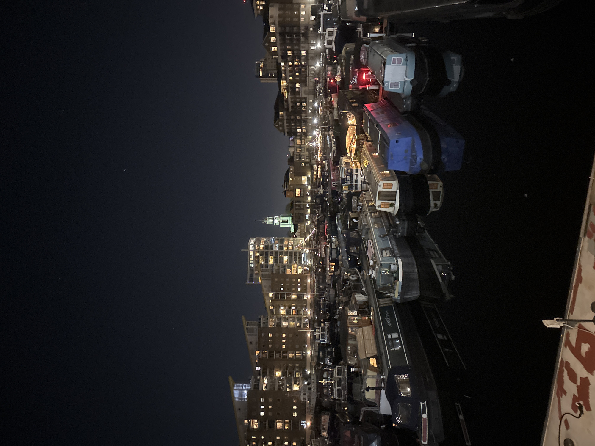 The image shows a nighttime scene along a canal in London, United Kingdom. A row of houseboats are docked along the canal, illuminated by interior lights. Buildings are visible on the opposite bank, with many illuminated windows. The sky is dark, with one visible star.
