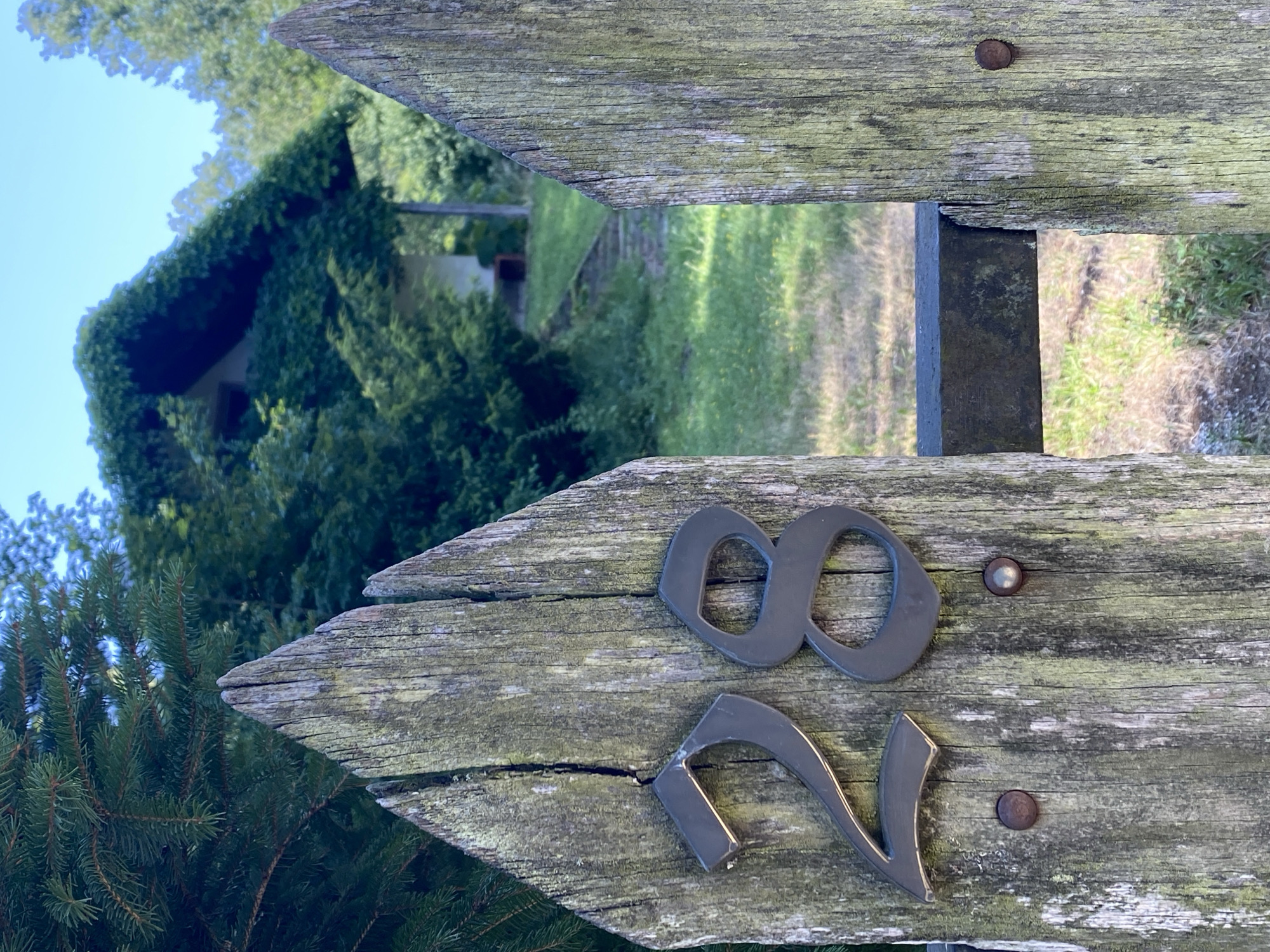 The image shows a weathered wooden gate with the numbers "28" attached to it. The gate is made of two pointed wooden posts, each showing signs of age and moss growth. The numbers are metallic and dark in color. In the background, a house covered in ivy is visible, with a pathway leading up to it. The setting appears to be a rural or suburban area with trees and green grass. The location is the city of Pressbaum, Austria.