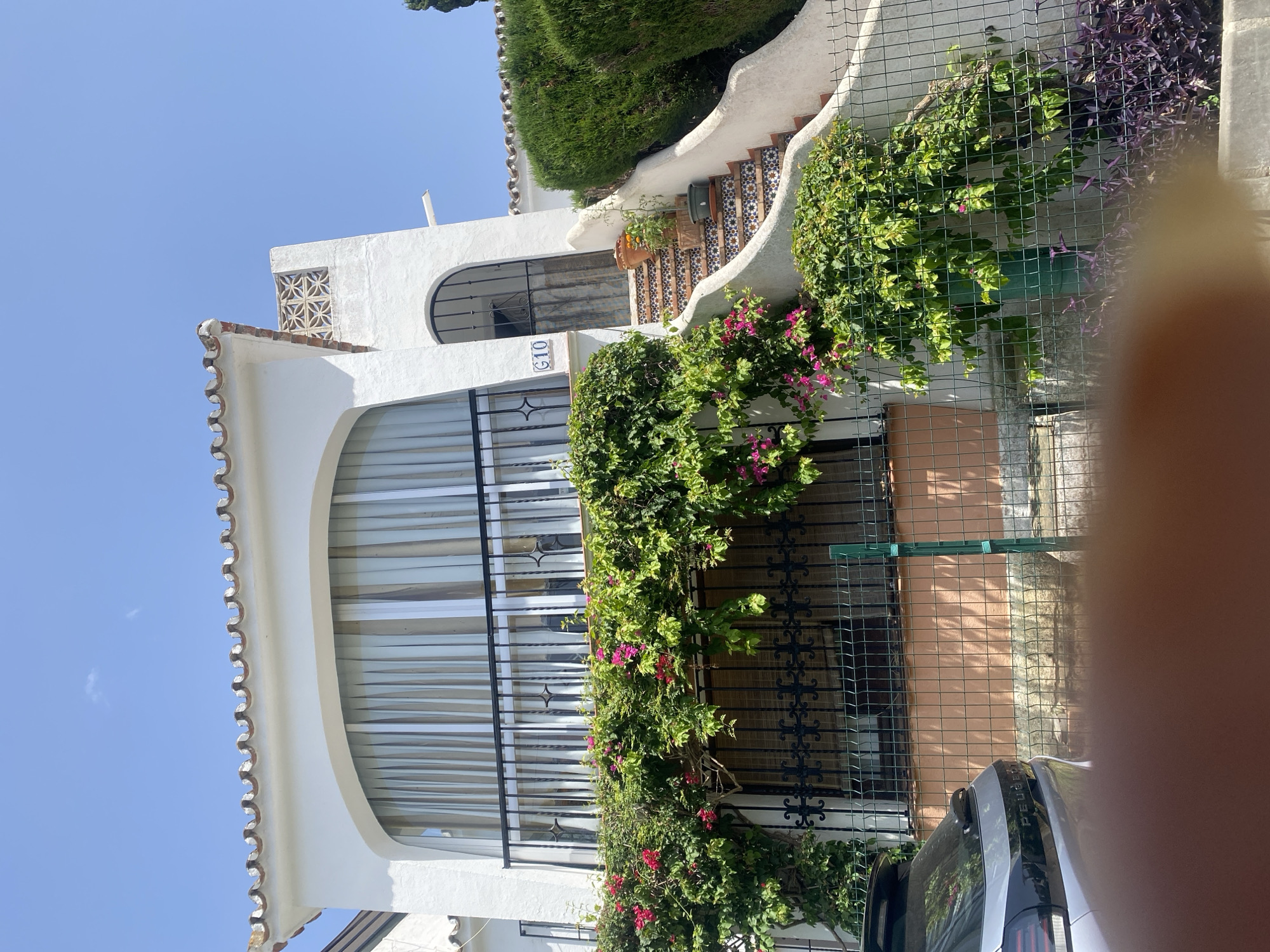 The image shows a white building with Spanish-style terracotta roof tiles, located in Paraíso-barronal, Spain. The building features a large arched window on the upper floor, partially covered by light-colored curtains, and a black metal railing along a balcony. Lush green plants with pink flowers are cascading down from the balcony, covering a portion of the lower facade. A set of stairs with decorative tile risers leads up to an arched doorway with a black metal gate. The number "G10" is visible on a blue and white tile near the doorway. A grey Peugeot car is parked in front of a wire fence that encloses the property. The environment appears sunny with a clear blue sky.