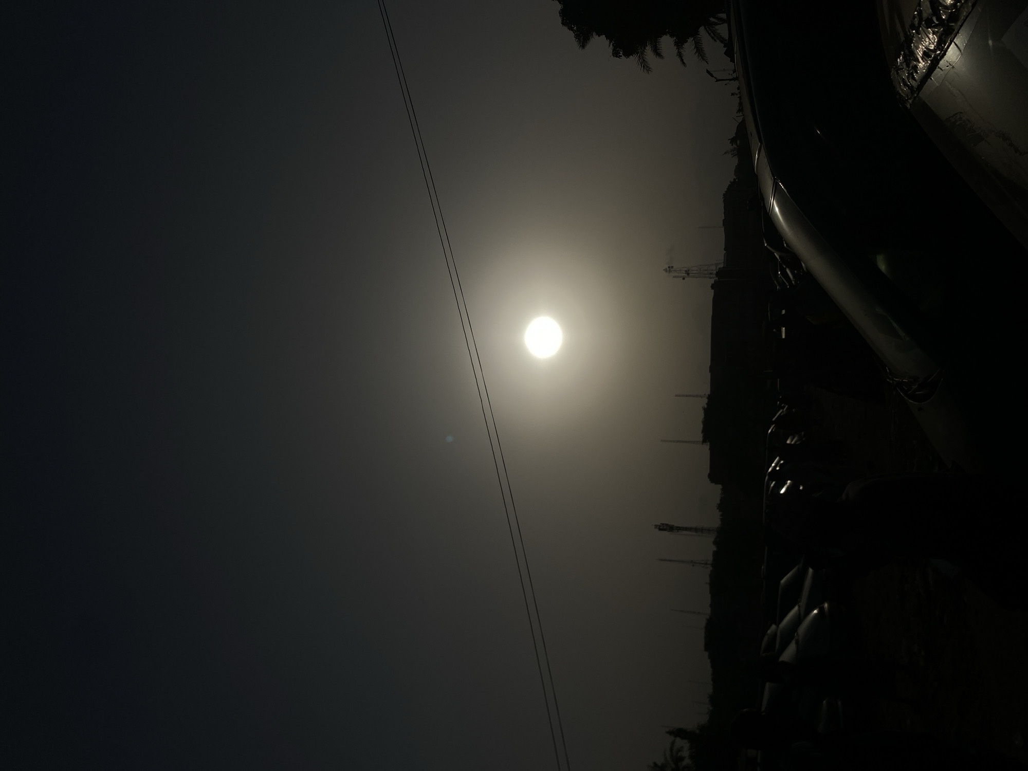 The image depicts an outdoor landscape under hazy, low-light conditions, suggesting either sunrise or sunset. A prominent, bright, circular light source, likely the sun, is positioned in the upper center-left of the frame, emitting a diffused glow across the sky. Two parallel power lines stretch diagonally across the upper portion of the sky, from the upper left towards the lower right, partially obscuring the light source.
Along the horizon on the right side of the image, faint silhouettes of various structures are visible, including what appear to be communication towers or industrial facilities, hinting at a semi-urban or industrial setting. Dark, indistinct shapes occupy the far-right foreground and edge, possibly belonging to a large vehicle, structure, or dense foliage, including the fronds of a palm tree visible in the upper right corner. The overall atmosphere is muted and somewhat somber due to the haze and low light. No specific activity or event is taking place, and there is no visible text. The weather appears hazy or foggy, contributing to the soft diffusion of the light. Given the context of Maiburiji, Nigeria, the scene reflects a typical hazy morning or evening in a region with both natural elements and infrastructure.