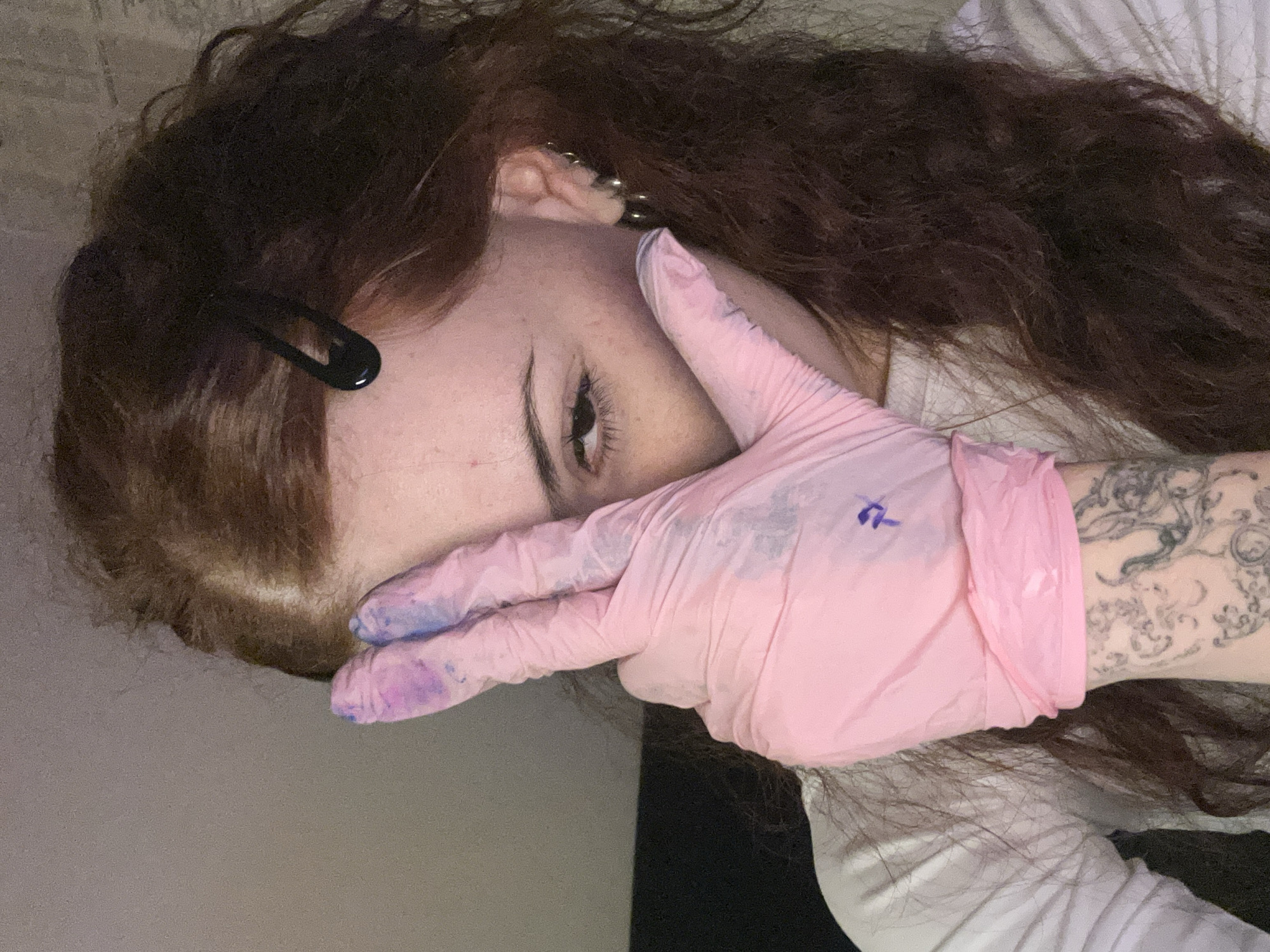 A person with long, wavy, auburn hair and fair skin is angled towards the camera, their face partially obscured by a pink gloved hand. The glove has blue smudges on the fingertips. A black bobby pin is visible in the person's hair near their forehead, and there are multiple earrings in their ear. The person's right arm is visible, adorned with a detailed tattoo. The person is wearing a white long-sleeved shirt. The background appears to be a plain wall or surface. There is no information provided indicating a location in Buenos Aires, Argentina.