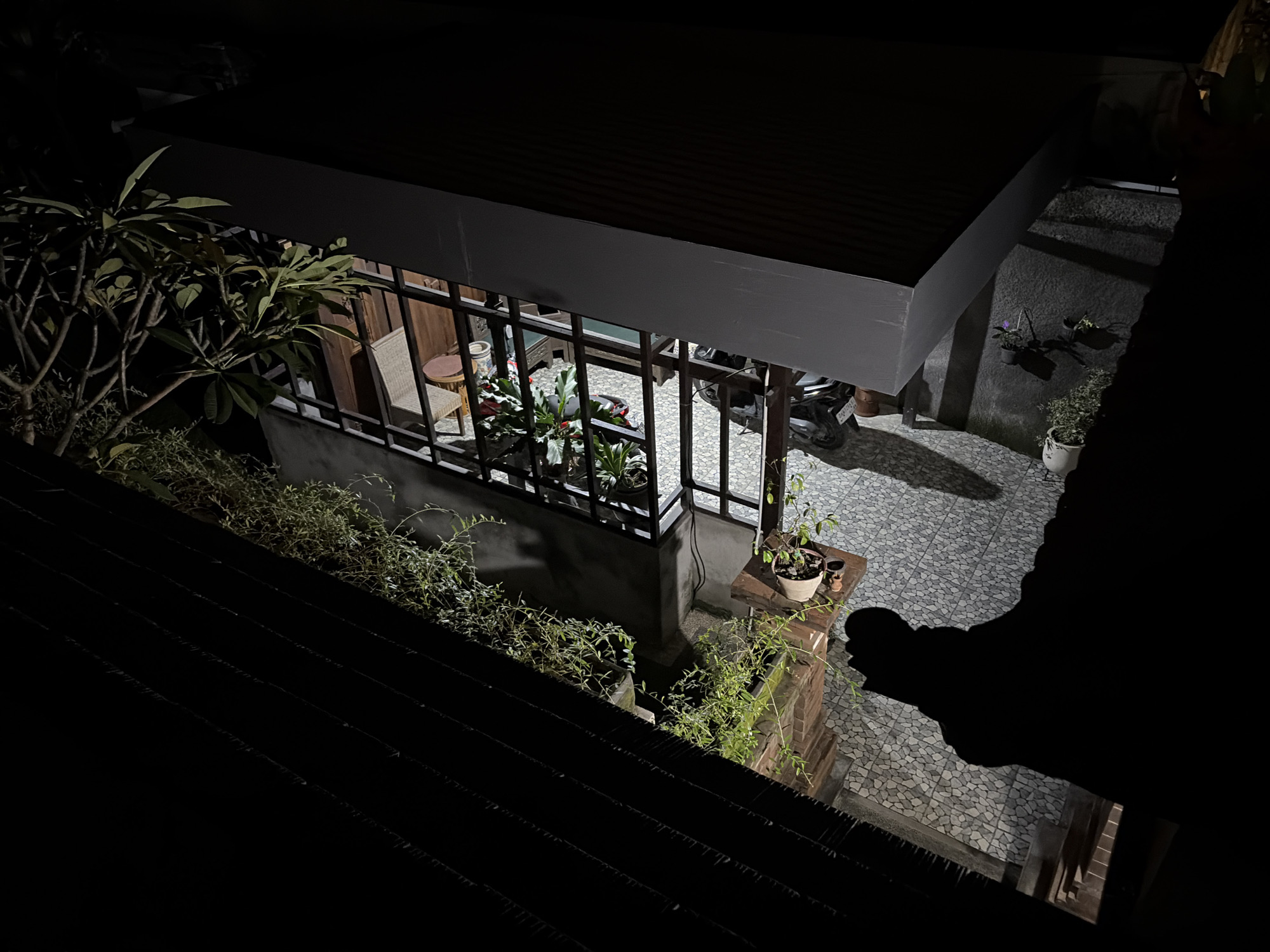 The image depicts a night scene in a courtyard area of Kuta Selatan, Indonesia, viewed from an elevated perspective. A dark, sloped roof occupies the foreground and lower portion of the frame. Beyond this, a covered outdoor patio area is visible, illuminated by artificial light. This patio features a patterned tiled floor and is enclosed by a dark metal railing. Inside the patio, there is a wicker chair and a small round table with a potted plant. Further within the patio, several other potted plants are arranged, and a motorcycle is parked. The walls of the structure behind the patio are dark and textured. To the left of the patio, a leafy tree with multiple branches is partially visible, its leaves illuminated by the ambient light. In the background, to the right of the patio, a wall is adorned with two small hanging potted plants and a larger potted plant on the ground. The overall scene suggests a residential or commercial outdoor space, typical of the Kuta Selatan region, at night. Shadows are prominent throughout the image due to the lighting conditions.