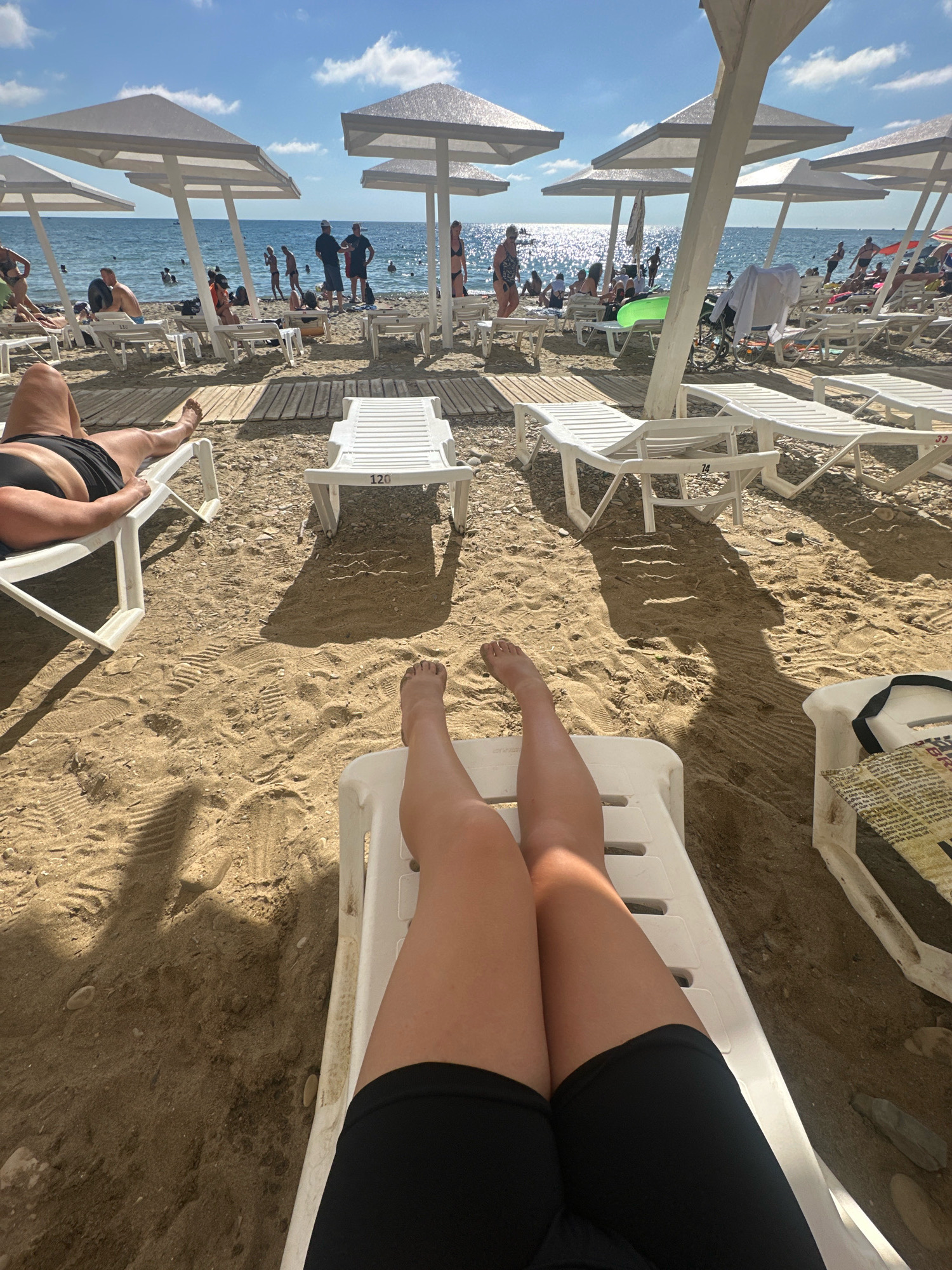 This is a first-person perspective shot of a beach in Lermontovo, Russia. The camera is positioned low, showing the legs of a person in black shorts and a black bikini bottom reclining on a white lounge chair. The sand is a light brown and appears to be packed and marked with footprints. In the foreground, more white lounge chairs are visible, some empty and some occupied by beachgoers. Beyond the chairs, the Black Sea stretches out under a clear blue sky with scattered white clouds. Numerous beach umbrellas, mostly white, provide shade for the loungers. A variety of people are scattered across the beach and in the water, some swimming and others relaxing on chairs or standing. The overall scene depicts a busy beach on a sunny day.