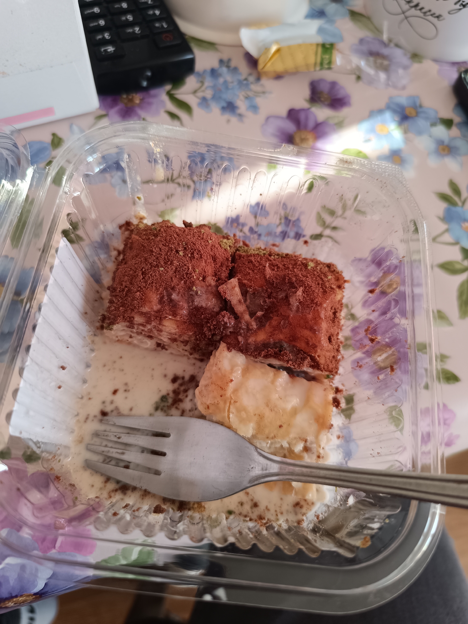 A clear plastic container on a floral-patterned tablecloth contains remnants of a dessert, partially eaten, alongside a silver fork. The dessert appears to be layered with a light-colored cream and topped with a dark brown powder, possibly cocoa. A portion of the dessert has been cut away, revealing the interior layers. The fork rests on the bottom of the container amidst the remaining cream sauce. In the background, out of focus, are objects including a remote control, a box, and a mug. The scene appears to be indoors. There is no information to establish that this is located in Sofia, Bulgaria.