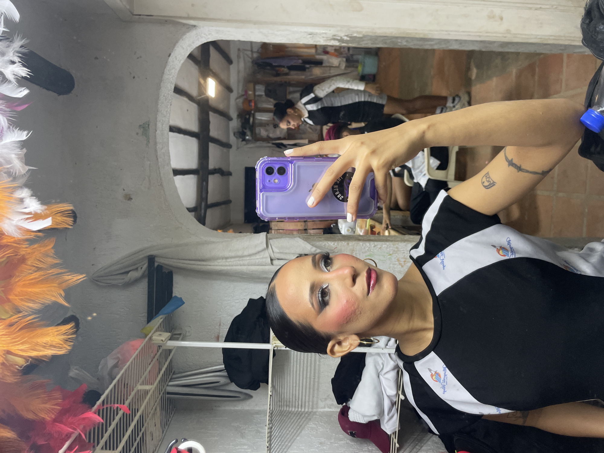 The image shows a young woman taking a selfie in what appears to be a backstage or dressing room area, possibly in Porlamar, Venezuela. She is wearing a black and white athletic top. Her hair is slicked back, and she has dramatic makeup on, including bright pink blush, silver eyeshadow, and dark lipstick. She is holding up a purple phone case in her right hand, which is covering her face. In the background, a mirror reflects another person wearing a similar athletic outfit and a cluttered environment with shelves, clothing, and brightly colored feathers hanging from the ceiling. The room has a rustic appearance with a textured wall and an arched doorway leading to another area with visible wooden beams and lighting. There are also some tattoos visible on the woman's arm.