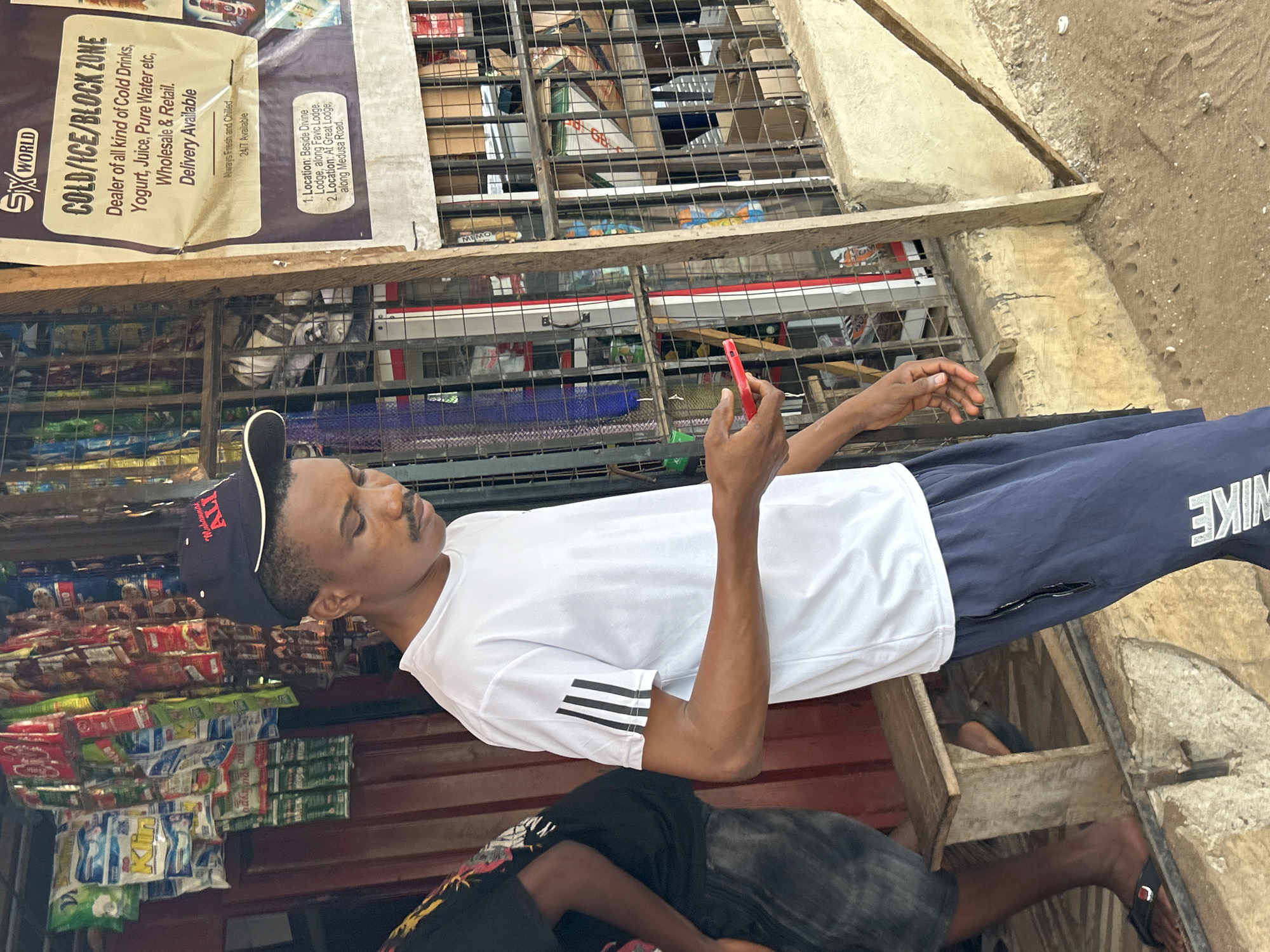 A young man wearing a white Adidas t-shirt with black stripes on the sleeves and navy blue Nike track pants with a zipper on the side, stands looking down at a red smartphone in his right hand. He is also wearing a dark baseball cap with "Muhammad Ali" embroidered in red. The man is standing in front of a stall or small shop that appears to sell drinks and snacks, with various packaged goods hanging from the ceiling and displayed behind a metal grate. A sign on a wooden post to the right of the man reads "SIX WORLD COLD/ICE/BLOCK ZONE" and lists the types of products sold. The location is identified as Ihiagwa, Nigeria. Another person, partially visible on the left, is wearing a black t-shirt and dark shorts. The ground is made of dirt and concrete.
