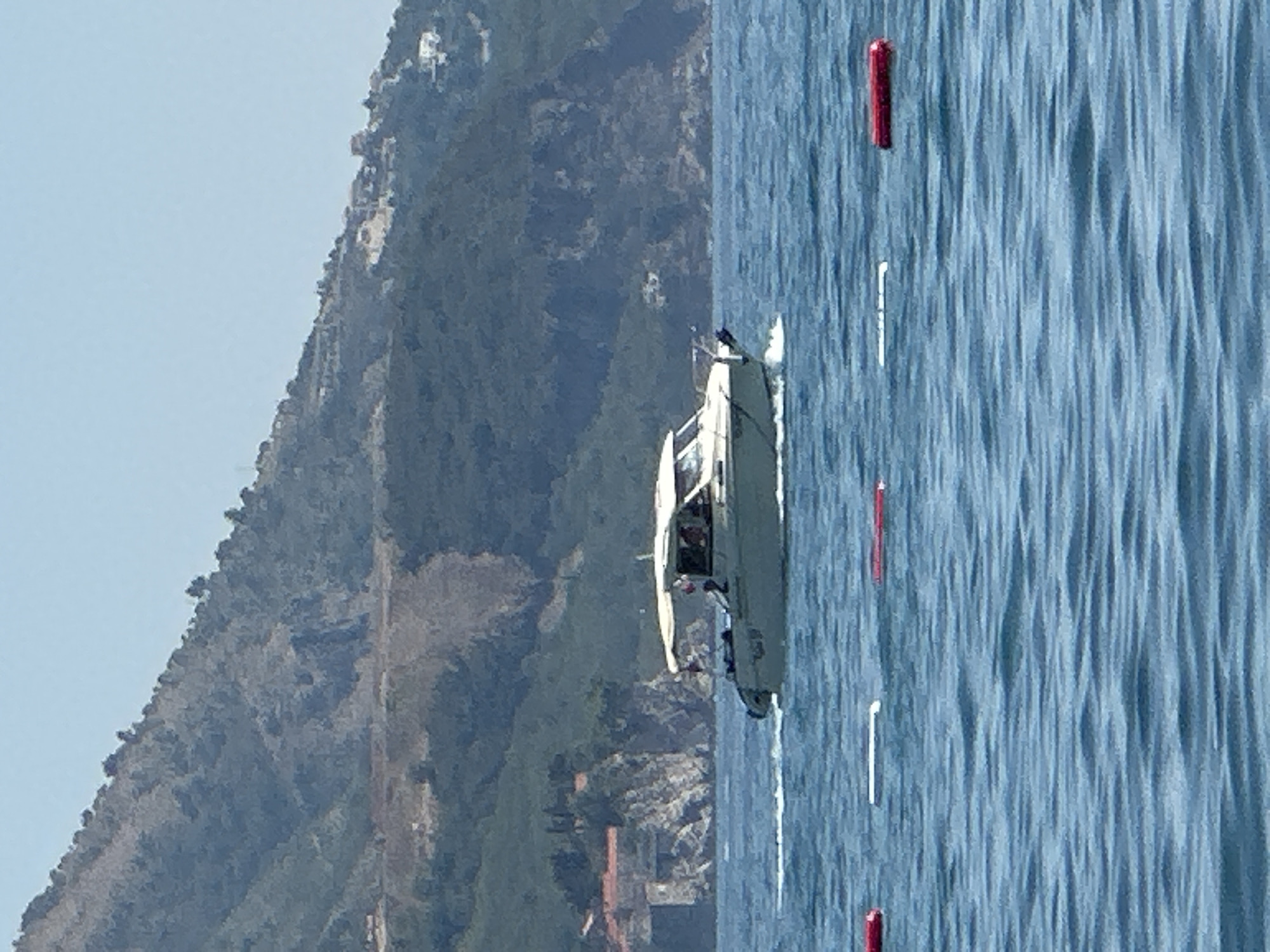 The image depicts a scenic view of the coastline near Boreti, Montenegro, under a clear blue sky. In the foreground, the deep blue sea is covered in gentle ripples, with several buoys marking a section of the water. These buoys include two red ones and one white one, arranged horizontally across the frame.
Further out, a motorboat navigates the water, leaving a white wake behind it. The boat appears to be light-colored, possibly white or cream, and has a canopy providing shade over the cockpit. Several individuals are visible inside the boat, suggesting it is in use for leisure or transport.
The background is dominated by a lush, green mountainside that slopes down towards the sea. The vegetation on the mountain is dense, with patches of rocky terrain visible. In some areas, structures are visible nestled on the slopes, indicating human habitation or development. These structures appear to be a mix of buildings with red-tiled roofs and lighter-colored houses. Rocky cliffs and outcroppings are also a prominent feature of the landscape, adding to the rugged beauty of the area. The overall impression is one of a serene coastal environment with a touch of human presence.