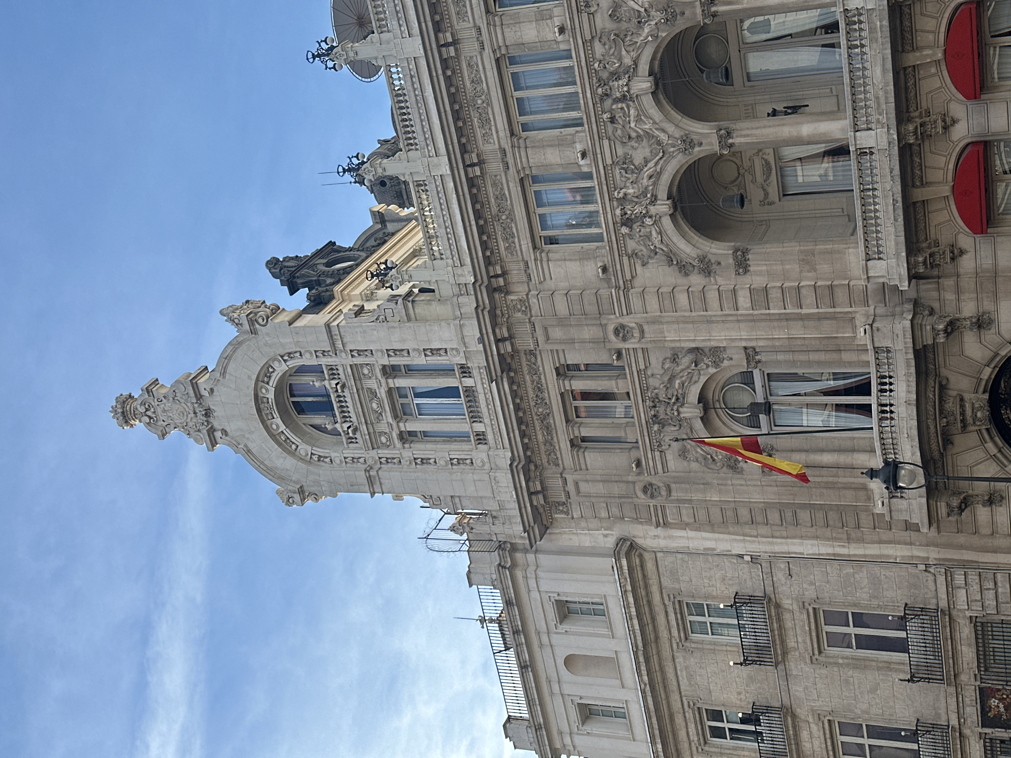 The image shows the top of a building in Madrid, Spain, taken from a low angle. The building's architecture is ornate with sculptural details, arched windows, and decorative cornices. A Spanish flag is visible hanging from a window. The sky is light blue with some wispy clouds.