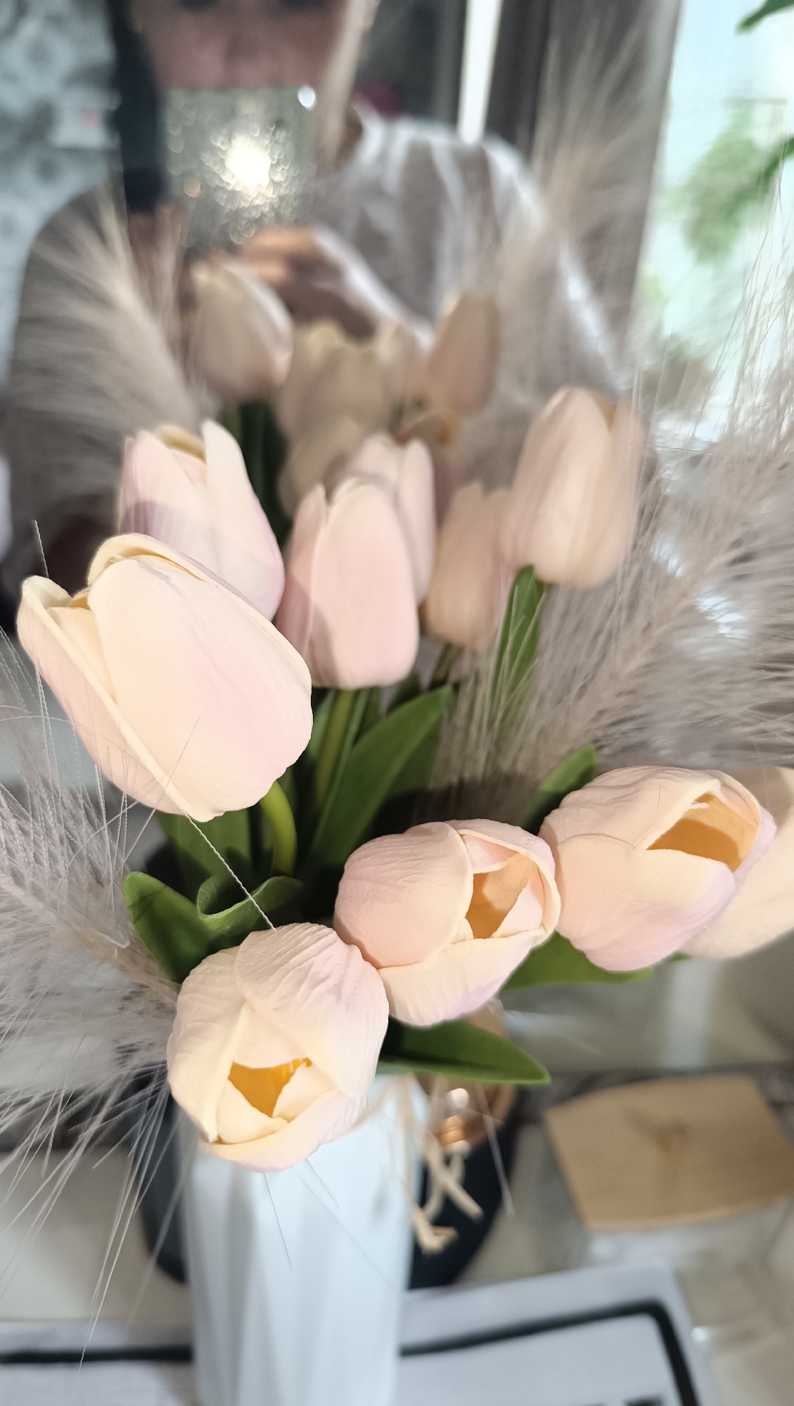 A close-up, vertical shot displays a bouquet of pale pink tulips arranged in a white vase. The tulips, with their delicate petals and yellow centers, are interspersed with fluffy, white decorative plumes. The background is blurred, showing a reflection in a mirror of a person wearing a white shirt, possibly taking the photo. The environment appears to be indoors, with hints of a bright window and a textured wall. The overall aesthetic is soft and decorative, suggesting a home or studio setting. The image does not contain any information about the city of Chaikovsky, Russia.