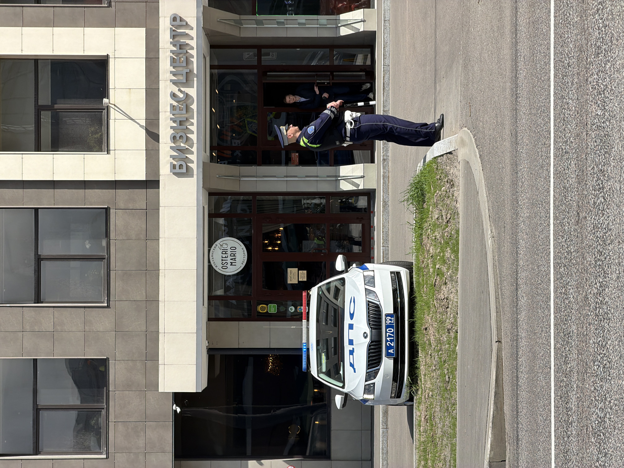 A police officer stands at attention outside a building in Unknown, Unknown. He is wearing a navy blue uniform with light blue accents, including a cap, jacket, and pants. A white police car with a blue and red light bar on top is parked behind him, facing the camera. The license plate number A 2170 99 is visible. The building has a sign in Cyrillic that reads "БИЗНЕС ЦЕНТР" above the entrance. Below the sign, there is a logo that says "OSTERIO MARIO". A man wearing a suit can be seen inside the building. The background includes the exterior of the building, a sidewalk, and a patch of grass. The image is shot in daylight with a clear sky.