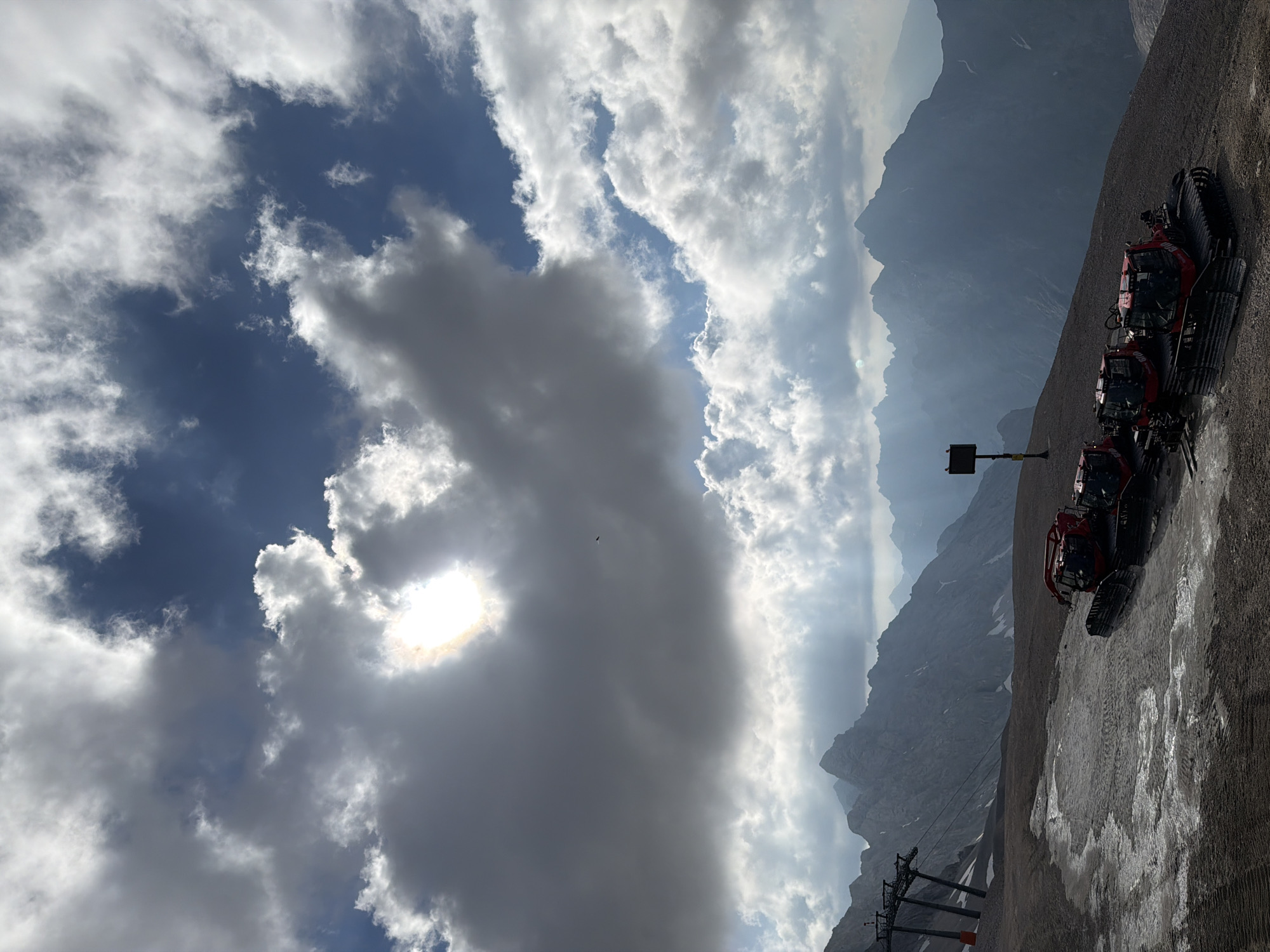 The image captures a scenic view of the Zugspitze, Germany's highest peak, located near Garmisch-Partenkirchen. The foreground shows a desolate, rocky terrain with patches of snow and ice, indicative of a high-altitude environment. Three red snow groomers, heavy machinery used for maintaining ski slopes, are parked on the ground. In the mid-ground, a ski lift structure stands tall, hinting at the presence of a ski resort. The background is dominated by a majestic mountain range, with jagged peaks shrouded in mist and illuminated by dramatic sunbeams breaking through an overcast sky. The sky itself is a canvas of dark and light clouds, with the sun partially obscured, creating a dynamic and atmospheric lighting effect. A single bird is visible in flight against the dramatic sky. The scene suggests a remote, mountainous landscape, likely a ski area in its off-season or during a time with limited snow cover at lower elevations.