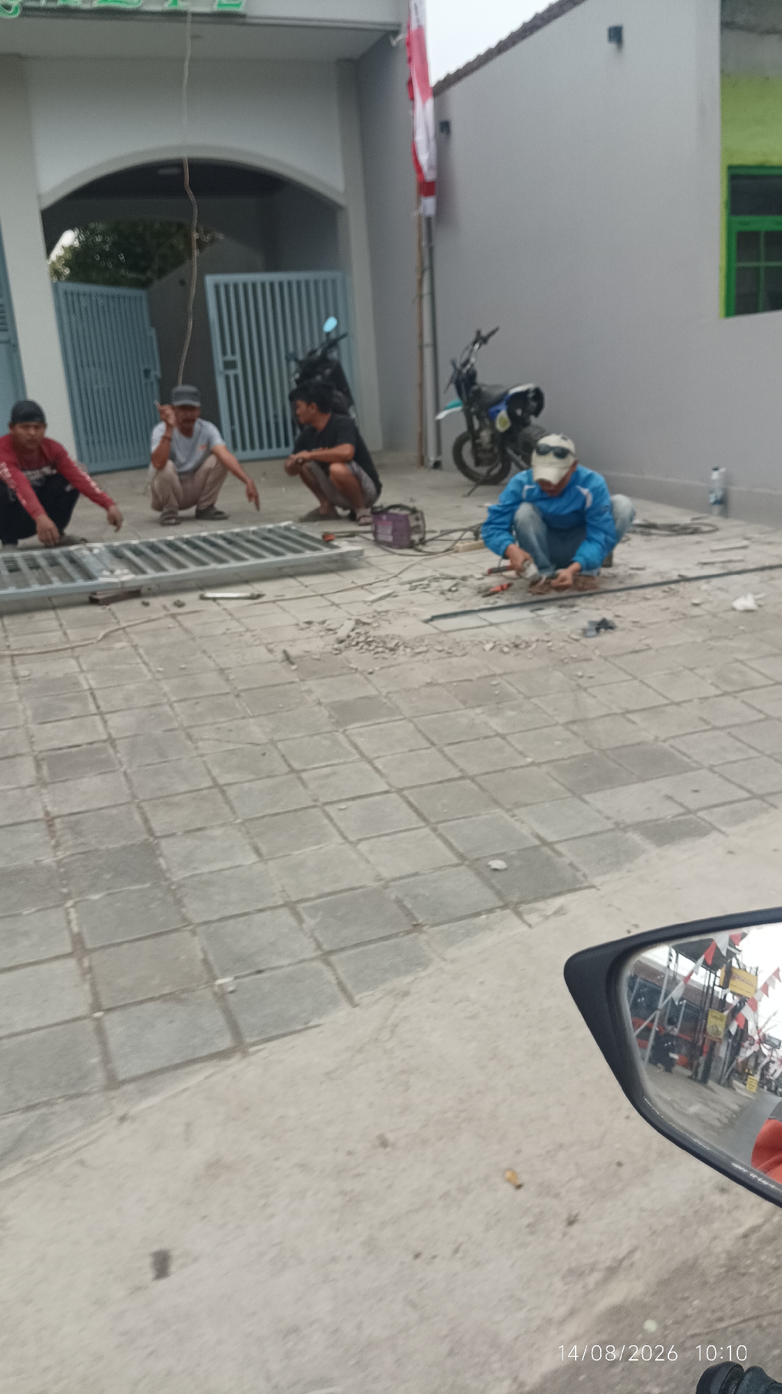 The image depicts an outdoor construction or repair scene in Tasikmalaya, Indonesia. Four individuals are present, actively engaged in work. Two men, one wearing a red long-sleeved shirt and the other a grey t-shirt and a cap, are positioned on the left side of the frame, appearing to be assembling or working with a metal grate. The third individual, wearing a black t-shirt, is also crouching on the left, near a welding machine and some debris. The fourth person, on the right, is wearing a blue jacket, white cap, and sunglasses, and appears to be working with tools on the ground.
The environment consists of a paved area with square tiles, a building with a light-colored facade and a dark roof in the background, and a blue gate. A motorcycle is visible behind the workers, and an Indonesian flag is prominently displayed on a pole to the right of the building entrance. Debris, including small stones and dust, is scattered on the ground around the workers, suggesting ongoing work. The scene is observed from a low vantage point, likely from the perspective of someone on a motorcycle, as indicated by the motorcycle's side mirror visible in the foreground, reflecting a street scene with other vehicles and flags. The timestamp "14/08/2026 10:10" is overlaid on the bottom right corner of the image.