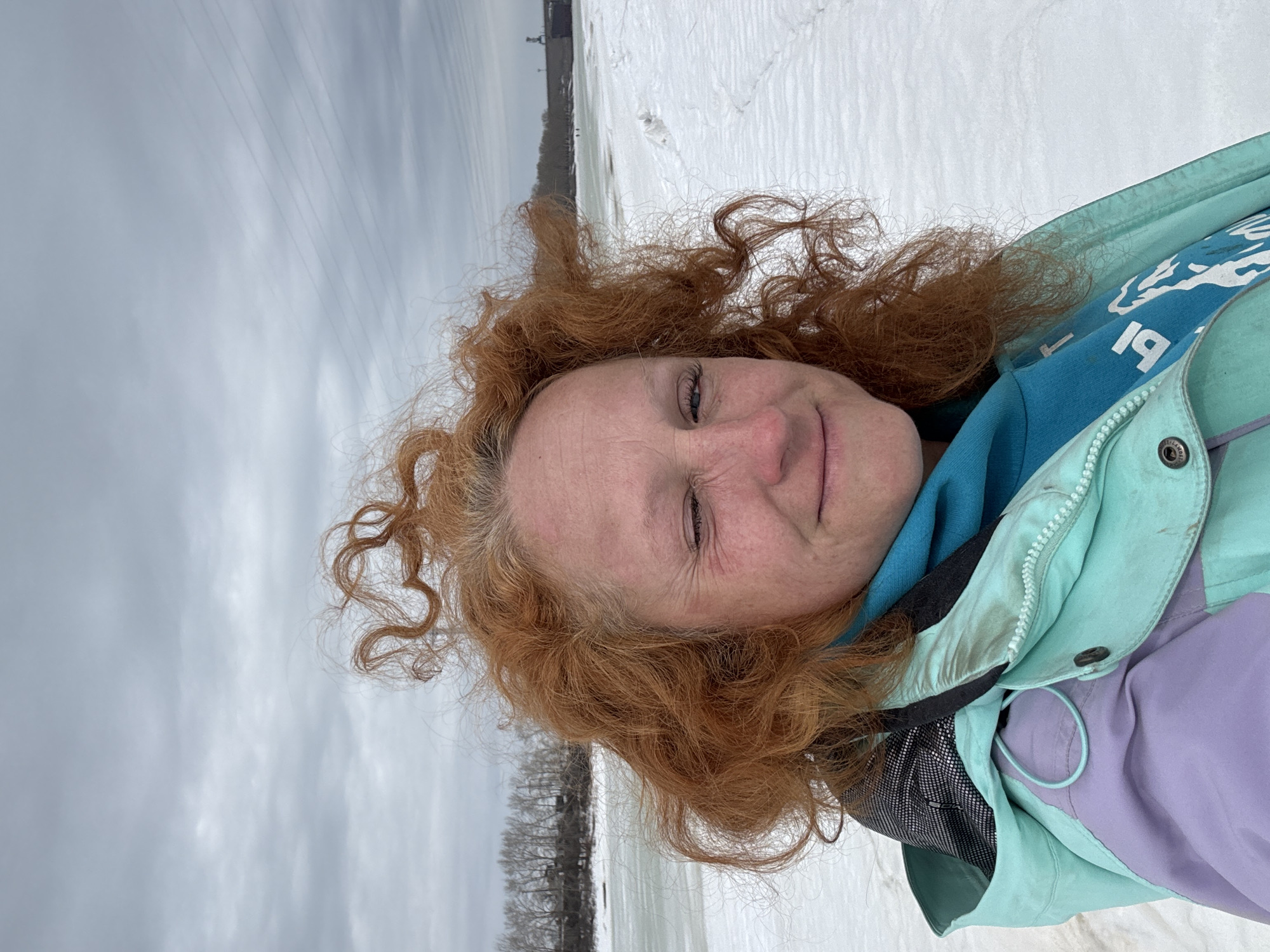 A fair-skinned person with red, curly hair, wearing a light turquoise and purple jacket over a blue hooded sweatshirt, is facing the camera and slightly smiling. The background shows a wintery landscape with snow-covered ground and an overcast sky. Bare trees are visible in the distance along with a few buildings. Based on the visible details, the location is suspected to be near a snowy, potentially lakeside area in the city of Chaikovsky, Russia.