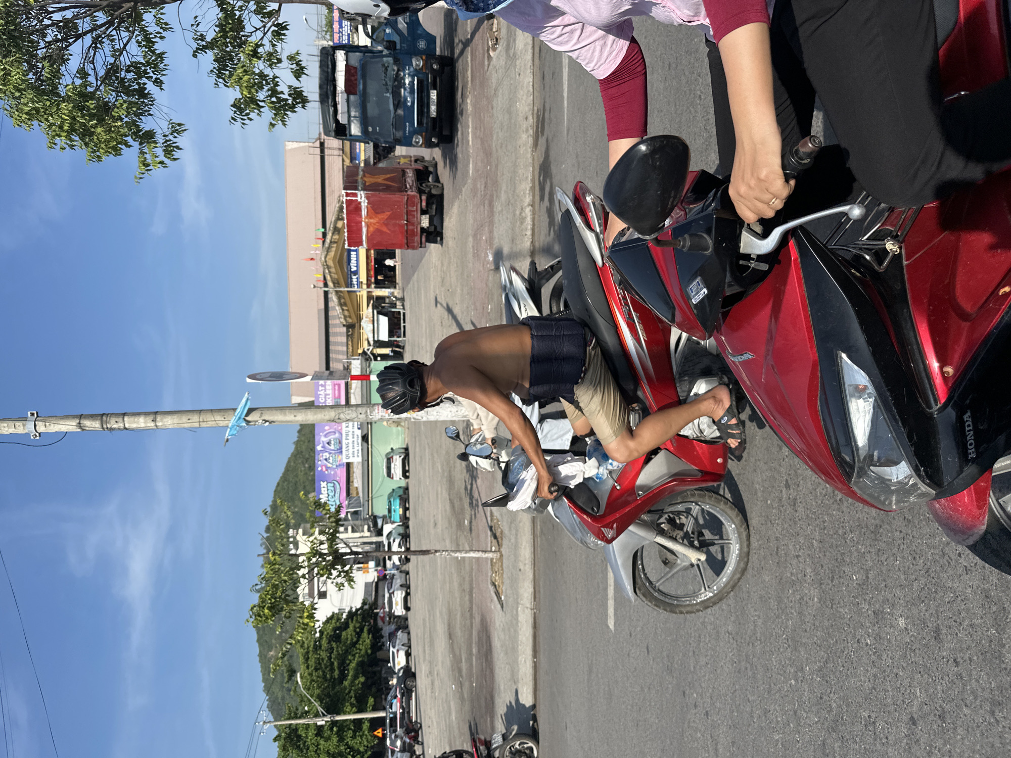 In Bac Nha Trang Ward, Vietnam, on a sunny day, two individuals are riding motorcycles on a paved road. The person in the foreground is wearing black pants and a pink long-sleeved shirt, gripping the handlebars of a red motorcycle. In the mid-ground, a shirtless person with a black helmet rides a red and silver motorcycle, wearing beige shorts and black flip-flops, and has a black back support strapped around their waist. Behind them, a blue truck with license plate 79C-092.48 and a red truck with a similar license plate are parked. In the background, several cars are visible in a parking lot, and a market building with yellow awnings stands under a blue sky with scattered clouds. Trees and a utility pole are also present in the scene.