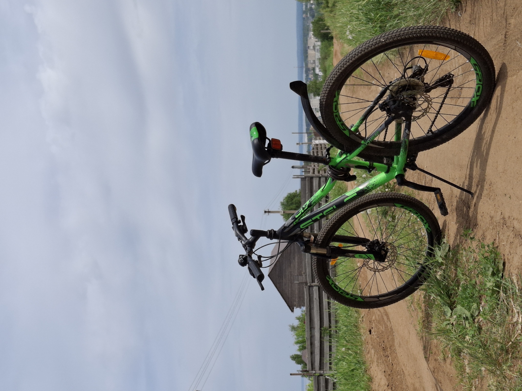 A green and black mountain bike is propped on its kickstand on a dirt path. The front wheel is facing the viewer. A small wooden building is visible in the background along with a wooden fence. In the distance, the cityscape of Chaikovsky, Russia can be seen with buildings and a body of water. The sky is overcast. Green grass and vegetation line the path.