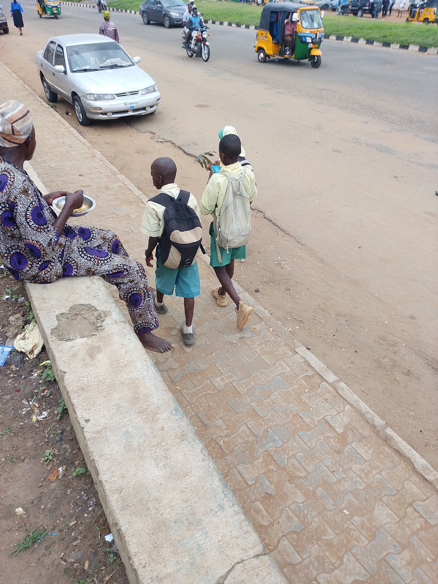 This media file captures a street scene in Oja Oba, Oko Erin, Nigeria. In the foreground, a person wearing a vibrantly patterned wrapper and headscarf sits on a concrete ledge, holding a small metal bowl containing food. To their right, two young boys in school uniforms walk along a paved sidewalk. The boy on the left wears a white shirt and blue shorts with a black and tan backpack, while the boy on the right wears a light yellow shirt and blue shorts, also carrying a backpack.
In the background, traffic moves along a dusty road. Several vehicles are visible, including a silver sedan, a yellow and green auto-rickshaw (Keke Napep), and a motorcycle with a rider. Other individuals can be seen in the distance, some walking or on motorcycles, indicating a bustling public area. The environment appears to be an urban or semi-urban setting with a mix of paved and dirt surfaces. The lighting suggests it is daytime, with overcast or diffused sunlight. There are no visible signs with text in this particular frame. The overall impression is a snapshot of everyday life and movement in a Nigerian community.