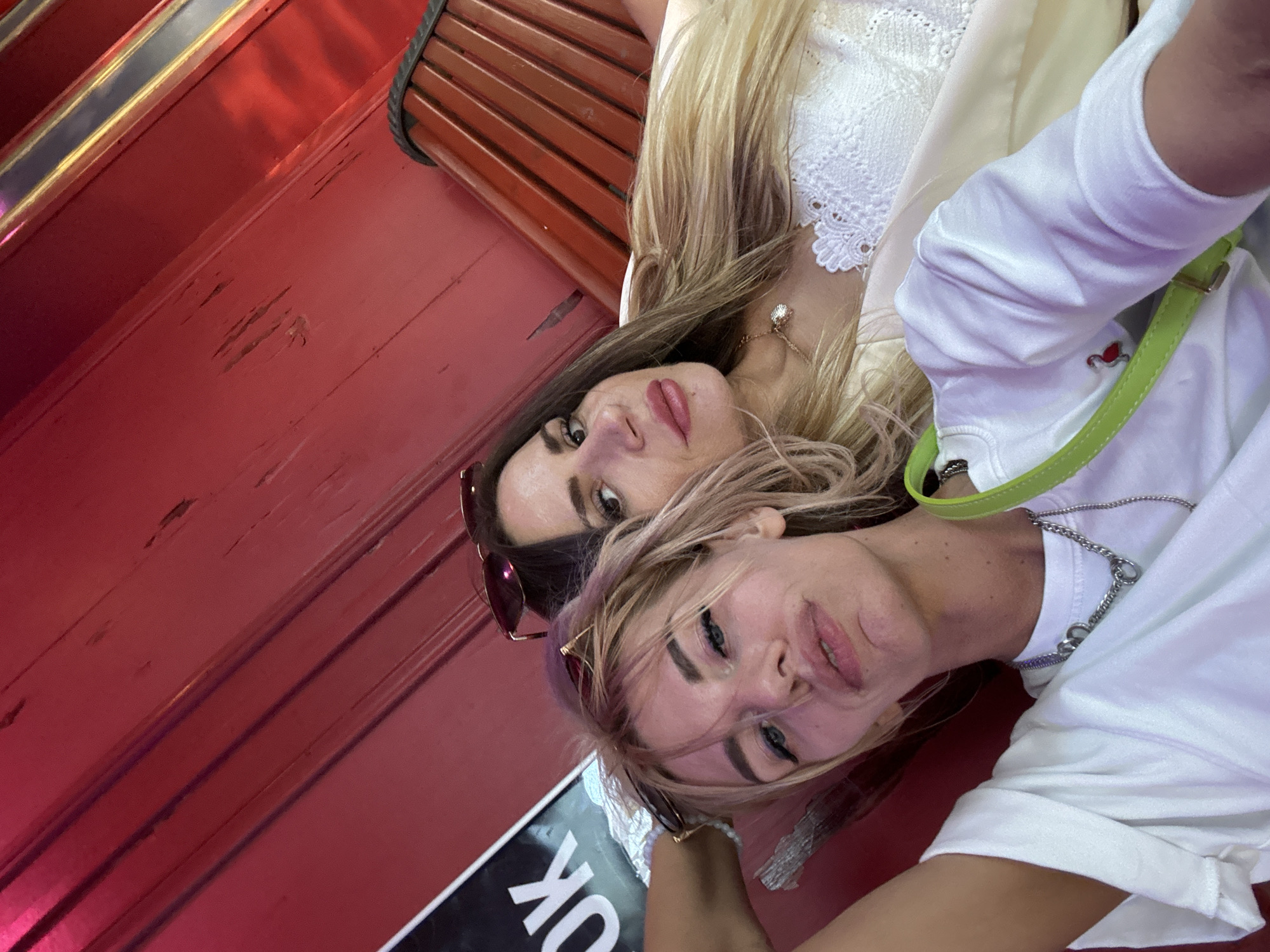 Two women are captured in a close-up, rotated selfie against a bright red, wooden background. The woman in the foreground on the left has blonde hair, is wearing a white top with a silver chain necklace, and a green purse strap is visible. She is making a puckered expression towards the camera. The woman behind her on the right has brown hair and is wearing sunglasses pushed up on her head, and a white lace top. She has a gentle smile. The background is a wall with horizontal wooden planks, painted a vibrant red. A black sign with white lettering, reading "OK", is partially visible above the blonde woman's head. The lighting is bright, casting a warm glow on the scene.