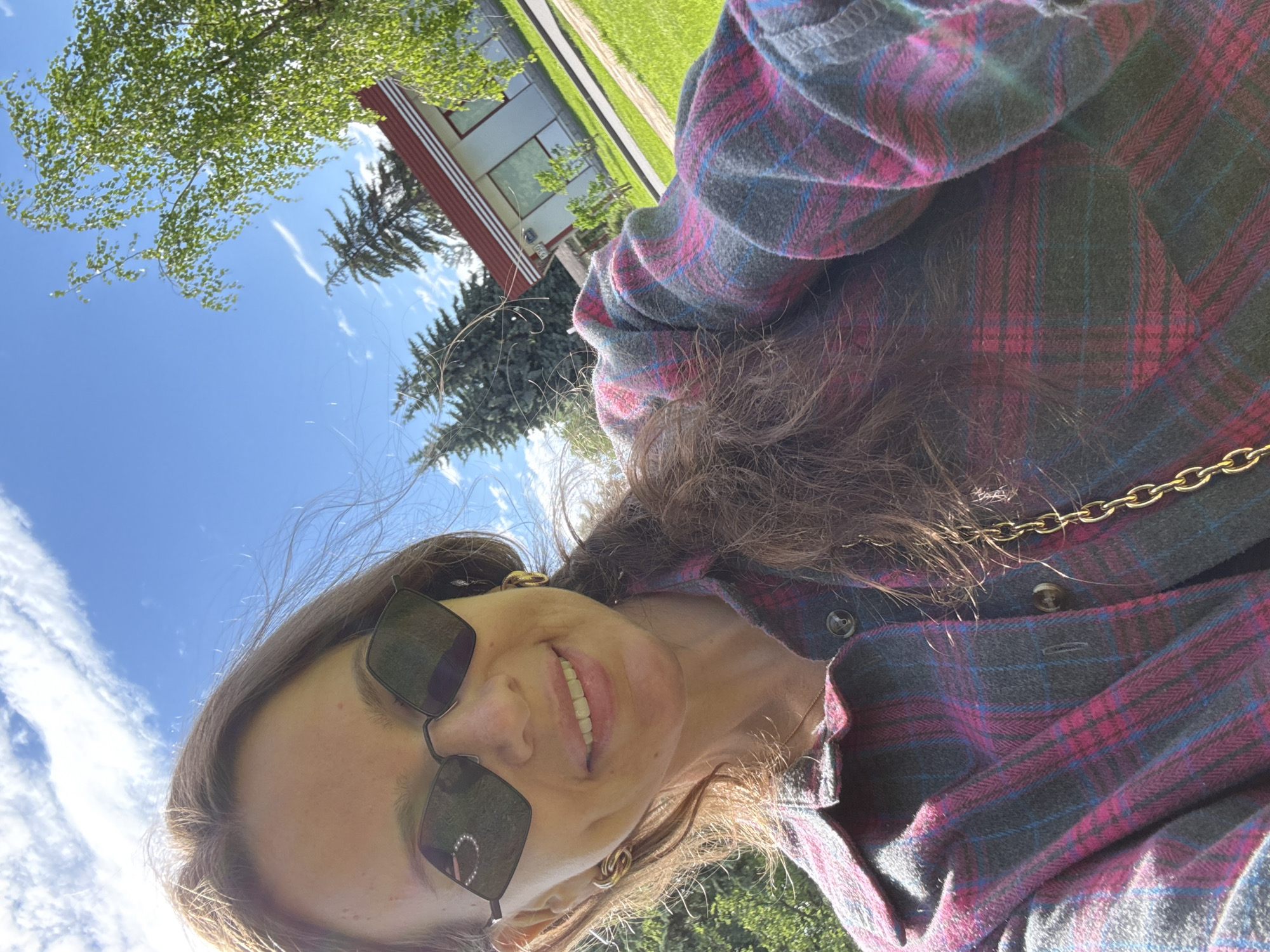 This is a close-up, selfie-style shot of a woman taken outdoors on a sunny day. The woman is wearing sunglasses with dark lenses and a plaid shirt in shades of pink, blue, and grey. She has long, dark brown hair that is slightly windblown. Her mouth is open in a smile, revealing her teeth. She is wearing a gold chain necklace. In the background, there is a blue sky with white clouds, green trees, and a building with a red roof and white trim. The overall impression is of a casual, pleasant outdoor activity. The lighting suggests it is daytime, likely late morning or early afternoon, given the brightness of the sun. The greenery and clear sky indicate fair weather. No visible text is present in the image.