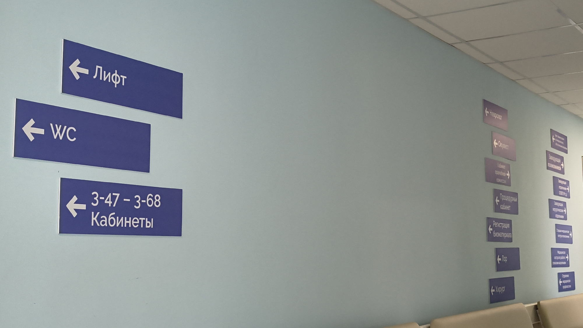 The image shows a hallway with several blue directional signs on a light blue wall. On the left side of the hallway, there are three signs stacked vertically. The top sign reads "Лифт" (Lift) with an arrow pointing left. Below it is a sign that reads "WC" with an arrow pointing left. The bottom sign on the left reads "3-47-3-68 Кабинеты" (3-47-3-68 Offices) with an arrow pointing left.
On the right side of the hallway, there are two columns of signs. The left column has signs reading "Невролог" (Neurologist) with an arrow pointing left, "Окулист" (Ophthalmologist) with an arrow pointing left, "Кабинет врачебной комиссии" (Physician's Commission Office) with an arrow pointing left, "Процедурный кабинет" (Procedure Room) with an arrow pointing left, "Регистрация биоматериала" (Biomaterial Registration) with an arrow pointing left, "ЛОР" (ENT) with an arrow pointing left, and "Хирург" (Surgeon) with an arrow pointing left.
The right column has signs with more text, including "Заведующая ПОЛИКЛИНИКИ" (Head of Polyclinic), "Заведующая" (Head), and other less legible text, all with arrows pointing left or down. The ceiling is visible at the top, with fluorescent lighting panels. In the foreground, a portion of beige upholstered seating can be seen. The environment appears to be inside a medical facility, possibly a polyclinic or hospital, given the nature of the signs. There are no people visible in the image. The location is stated to be in Mikhaylovka, Russia.