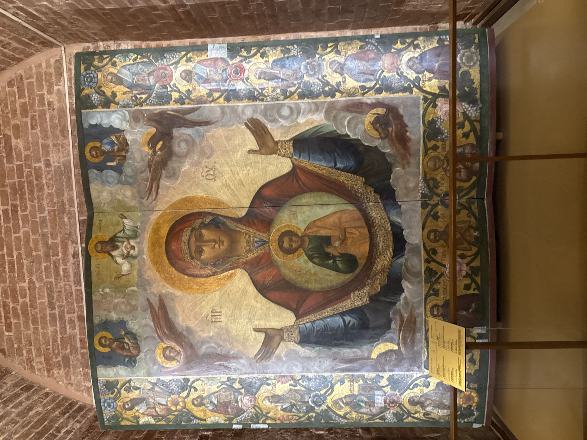 The image depicts a large Orthodox icon, likely the "Sign" icon of the Theotokos (Virgin Mary), displayed within the interior of a building with brick-vaulted ceilings, suggesting an older structure, such as a church.
The icon features Mary with her hands raised in orans (prayer), and a medallion on her chest containing an image of the Christ Child. The icon is elaborately painted, with figures of angels and saints surrounding the central image of Mary. The edges are decorated with further depictions of saints and floral patterns. The overall color scheme is rich, with gold, red, and blue dominating.
The building is most likely located in Moscow, Russia, due to the traditional style of the religious icon, the architectural style of the building.
