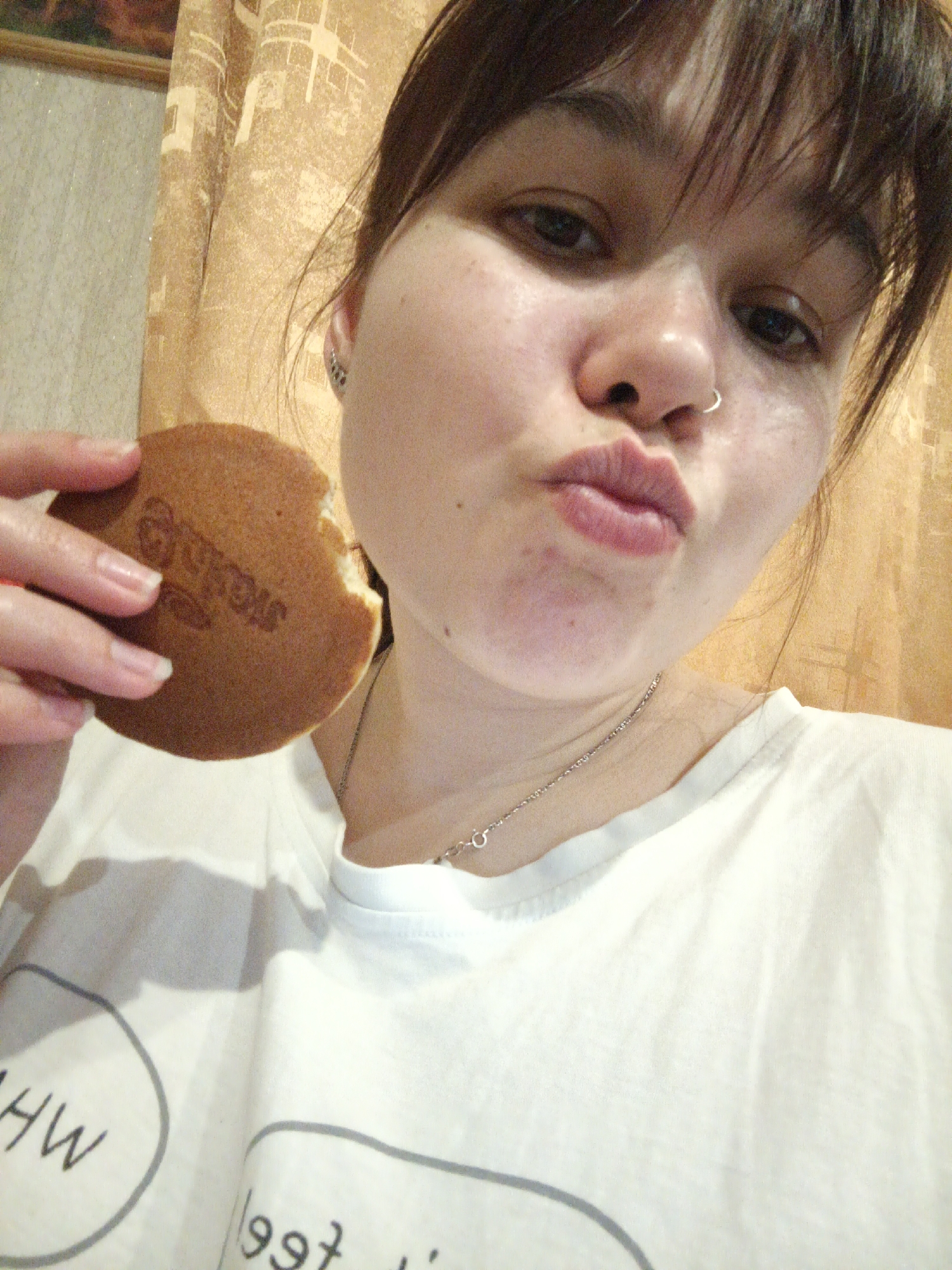 A young woman is taking a selfie indoors. She is fair-skinned with dark hair and bangs, and she has a nose ring. She is wearing a white T-shirt and a silver necklace. She is holding a round pastry with a bite taken out of it, with an inscription in a foreign language on the top. Her lips are pursed in a kiss. A curtain hangs in the background. The location is Chaikovsky, Russia.