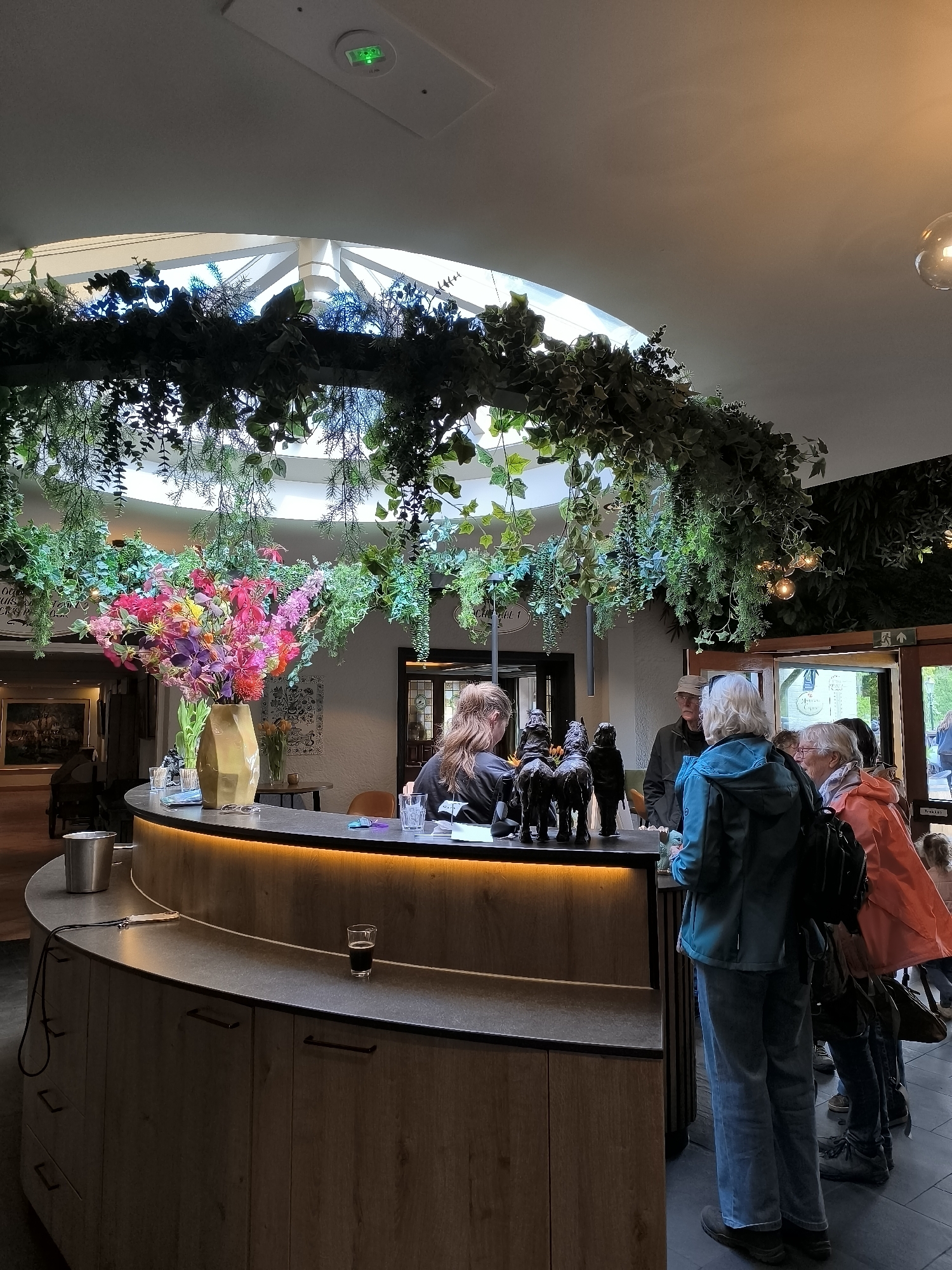 Here is a detailed description of the image:
**Scene:**
The image captures a bustling scene at the reception desk inside a building. The space has a unique architectural design featuring a high, circular opening in the ceiling, adorned with an elaborate arrangement of greenery and hanging plants.
**Environment:**
The interior design has a blend of natural and modern elements. The reception desk has a curved shape, constructed with light-toned wood paneling. A soft, warm light strip runs along the top edge of the desk, providing illumination.
**People:**
A group of people is gathered at the reception desk, seemingly waiting to be attended to. An individual in a blue jacket stands prominently in the foreground, while others are visible in the background. The receptionist, with her back to the camera, appears to be assisting customers. A man is visible with a hat waiting in line.
**Objects:**
On the reception desk, there are various items, including small sculptures of horses or animals, a glass with a dark liquid, and other decorative objects. The floral arrangement adds a pop of color to the scene.
**Location:**
While I do not have explicit confirmation of the location being in Castricum, Netherlands, some visual details might suggest a public or commercial space with a modern yet nature-inspired design aesthetic. There is also signage that says "Tickets" at the end of the line.