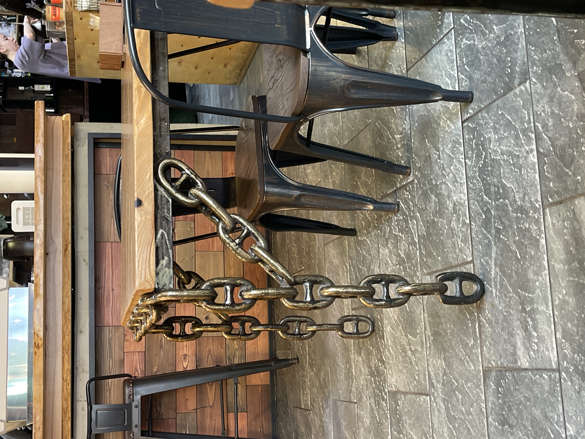 This image shows a close-up of heavy, brass-colored anchor chains lying on a stone-tiled floor. The chains are thick and appear to be decorative, perhaps used as a nautical-themed accent in a public space. They are arranged in a somewhat haphazard, draped fashion.
In the background, parts of wooden tables and dark metal chairs are visible, suggesting an indoor setting like a restaurant or cafe. A person's arm and shoulder can be seen in the upper left, wearing a light purple shirt. They appear to be interacting with something on a shelf or counter, though the details are unclear. The lighting suggests it is daytime, with natural light illuminating the scene.
The overall impression is one of a casual, possibly rustic or industrial-chic environment, with a nautical or maritime influence due to the prominent display of the anchor chains. The stone tiles have a marbled or textured appearance, and the wood elements appear to be light-colored. No text is clearly legible in the image. The context provided indicates the location is Zelenogradsk, Russia, which might align with a coastal town where nautical themes are common.