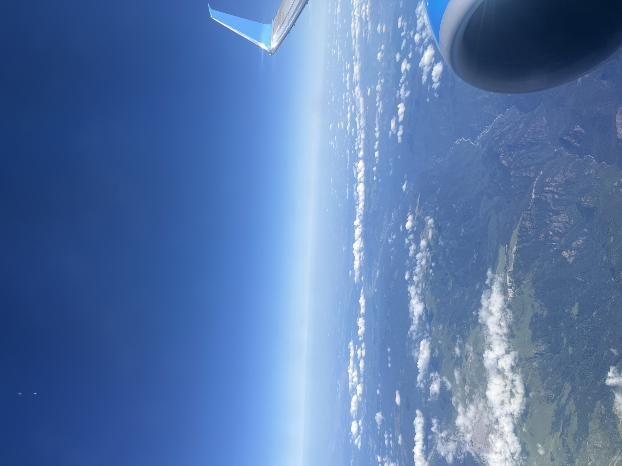The image is an aerial view from an airplane, looking out over a mountainous landscape and the sky. The upper portion of the image is a clear, bright blue sky with a faint haze near the horizon. In the far upper left corner of the sky, there are two small, bright, white dots. A portion of the airplane's wing, with a blue tip, is visible on the right side of the frame. Below the sky, a vast expanse of green, forested mountains stretches out, dotted with patches of white clouds. A winding river or stream can be seen in the valleys between the mountains. The foreground on the right shows part of the airplane's engine cowling, which is dark gray with a blue accent. The image does not provide any visual information that could pinpoint a location within the city of Sirius, Russia, or any specific details about people or actions.