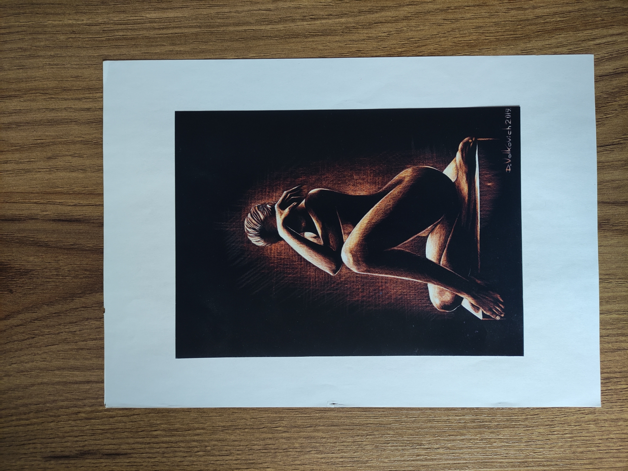 The image is a photograph of a print or artwork depicting a nude figure in a dramatic pose. The figure is lying on their side, with their legs bent and arms raised to their head. The lighting is focused on the figure, creating strong contrast and shadows. The background is dark, possibly black or deep brown. The artwork is mounted on a white card or paper, which is then placed on a wooden surface. The signature "D.Volkovich 2019" is seen written vertically on the right side of the artwork. The photo is taken indoors, likely on a table, with a focus on the artwork itself. There is no information in the image to suggest its location is in Tbilisi, Georgia.