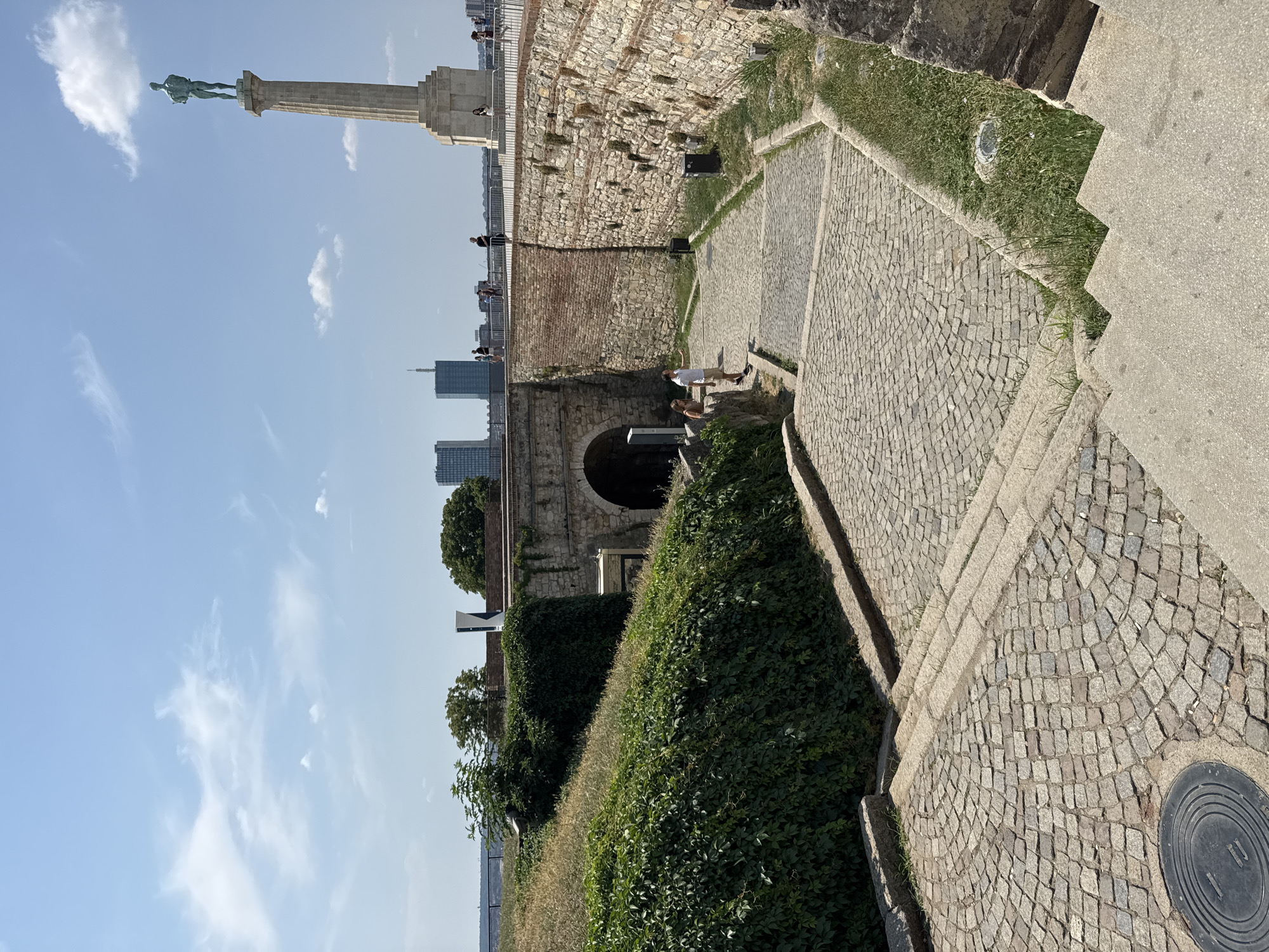 The image depicts a view of Kalemegdan Fortress in Belgrade, Serbia, on a sunny day. In the foreground, a cobblestone path curves downwards, leading towards an arched entryway built into a stone wall. To the left, a grassy slope covered in dense green ivy ascends. To the right, the stone wall continues, its rough-hewn blocks interspersed with brickwork.
Further into the scene, two individuals, a man and a woman, are standing on the cobblestone path, facing the arched entryway. The man, wearing a white t-shirt and shorts, appears to be gesturing towards something. The woman, in a light-colored dress, stands beside him.
Beyond the archway, a wider paved area leads to a walkway with a metal railing. In the distance, a prominent monument, the Victor Monument, stands tall on a pedestal. Behind it, a city skyline is visible, featuring modern high-rise buildings. Several people are scattered along the walkway, observing the sights. The sky above is a clear blue with scattered white clouds. The overall scene is one of historical architecture blended with a modern urban landscape, set against a bright summer day.
