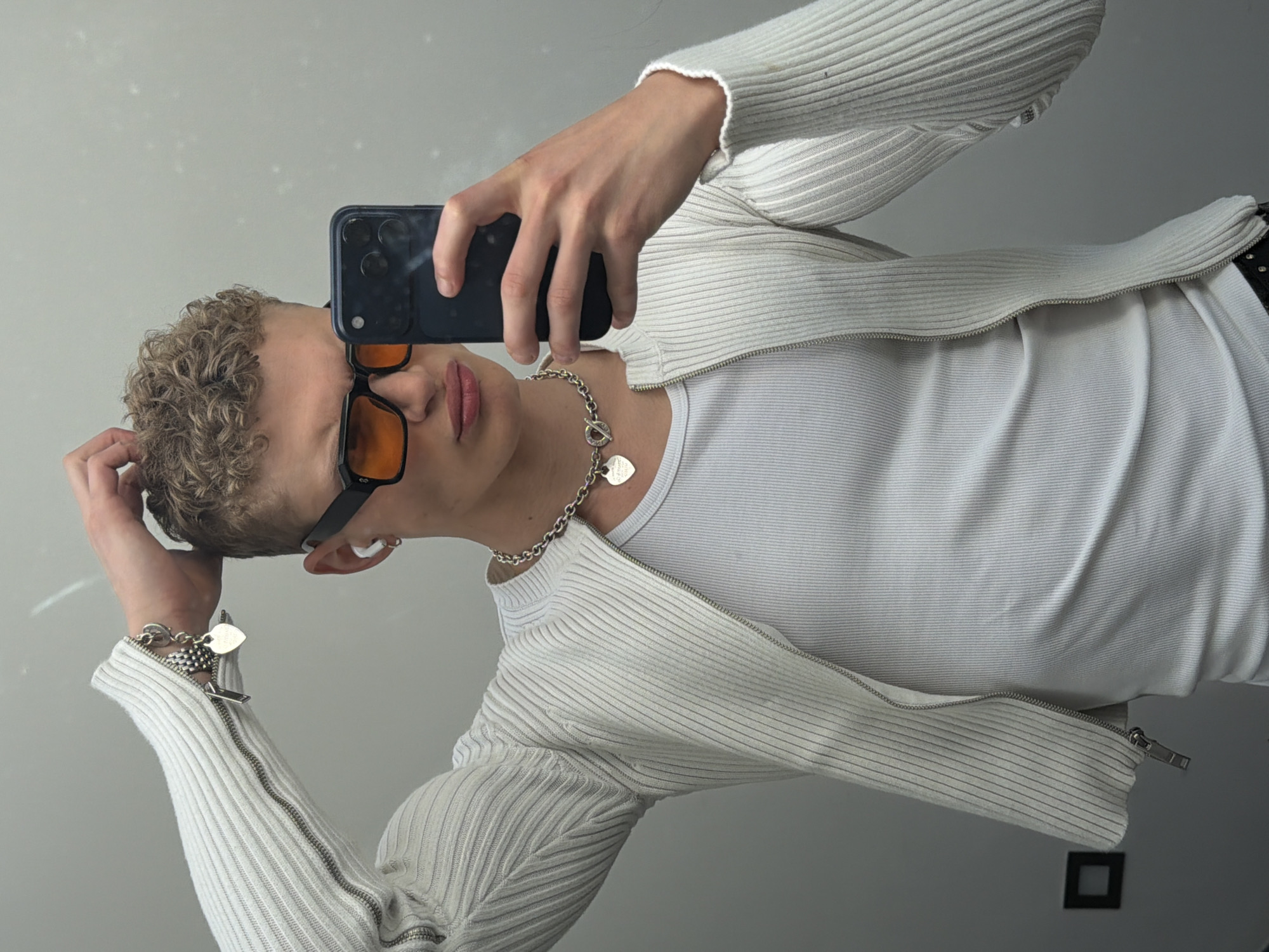 A young man is taking a selfie. He is wearing a white ribbed cardigan over a white t-shirt, black-framed sunglasses with amber lenses, and a silver chain with a heart pendant. He also has a silver bracelet on his left wrist. His hair is short and curly. The background is a plain light grey wall. A small black square (likely a light switch cover) is visible in the lower right corner. The location is likely a home interior in an unknown area of London, United Kingdom.