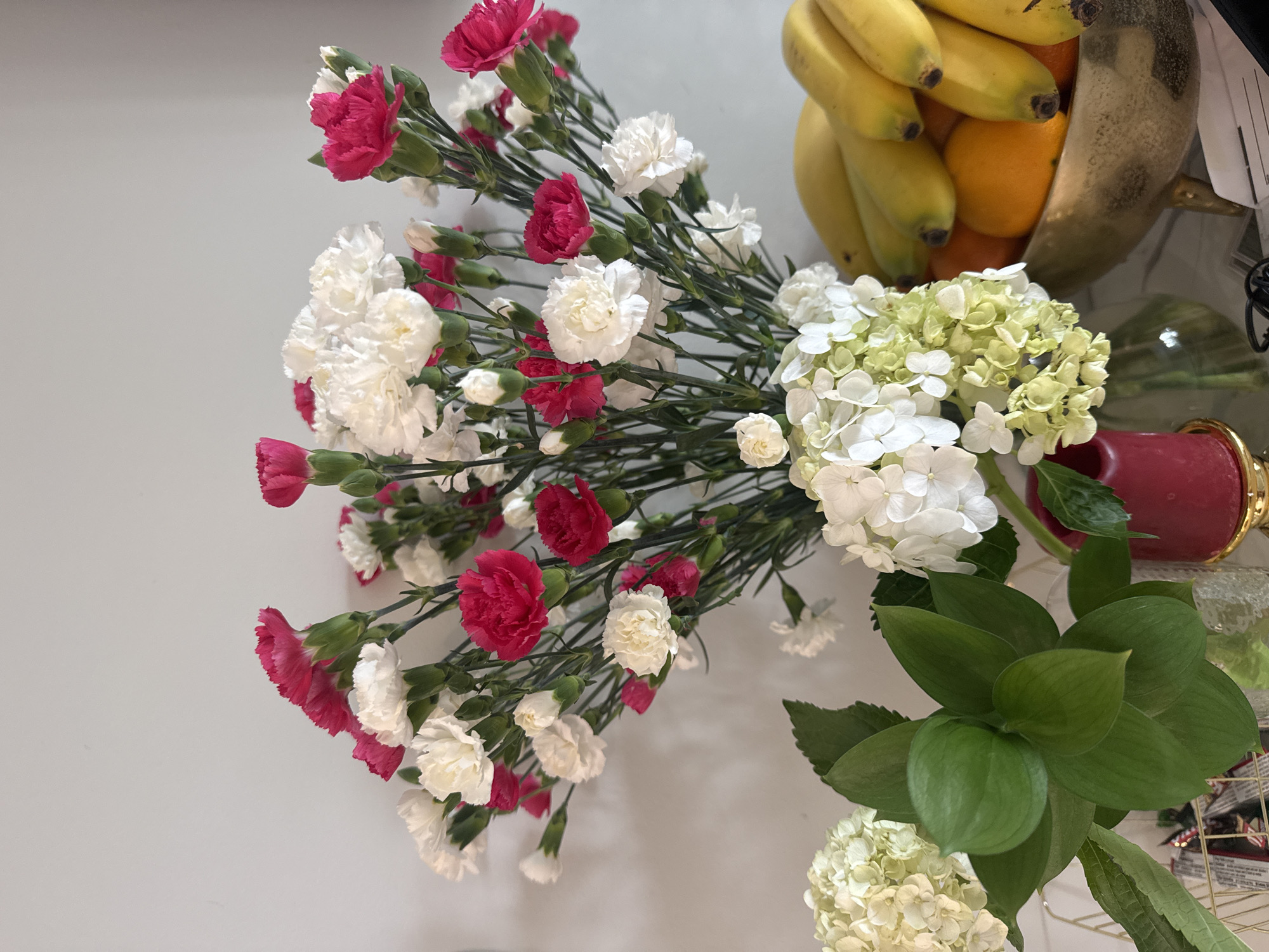 A still life arrangement features a bouquet of pink and white carnations in a clear vase. To the left of the carnations, a green leafy plant is displayed in a clear vase, next to a red candle in a gold holder. To the right of the carnations and leafy plant, a gold bowl filled with bananas and oranges sits on a white surface, possibly a countertop or table. The background is a plain, light-colored wall. The scene is indoors and appears to be in a domestic setting.