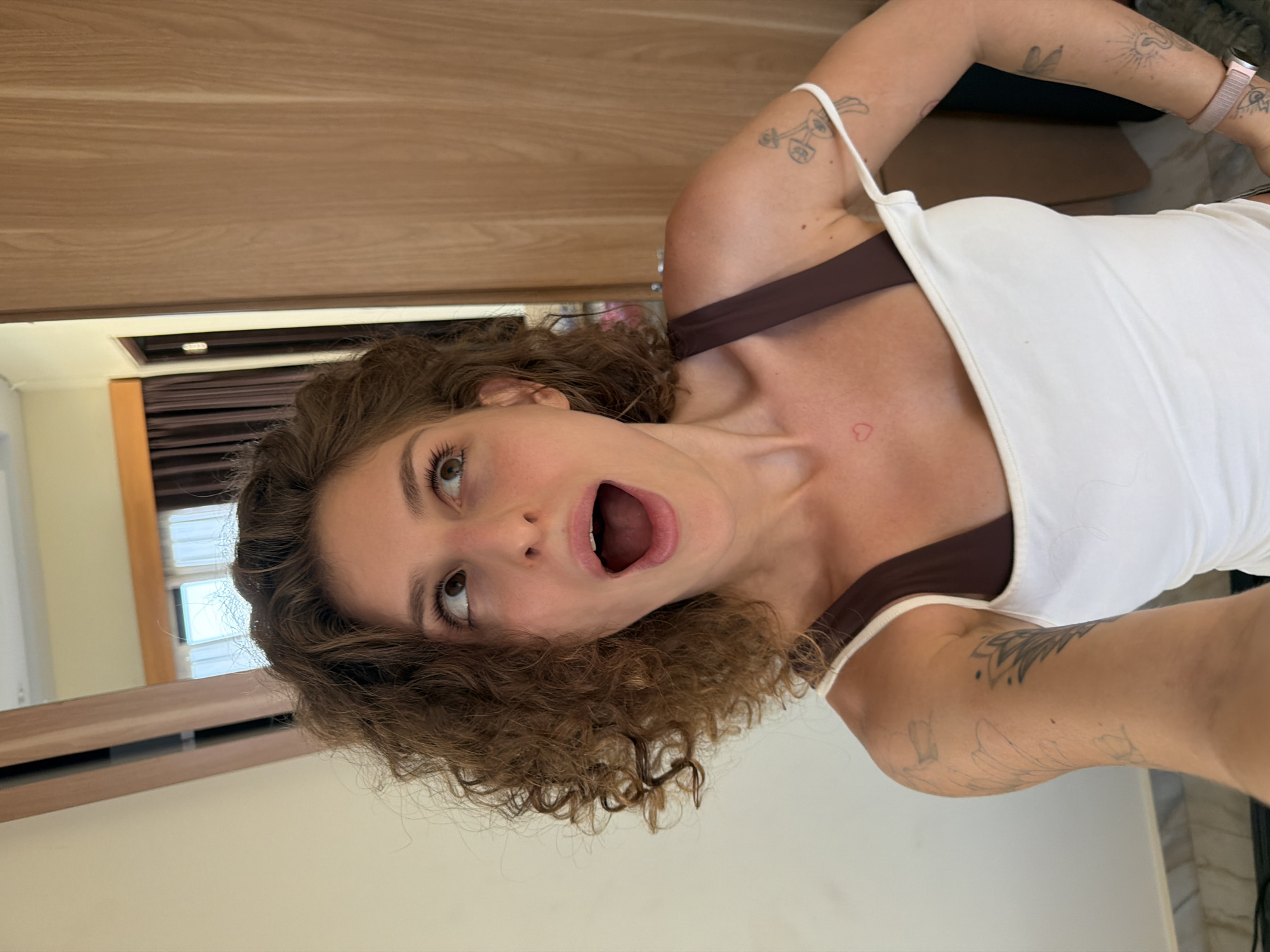A young woman with curly brown hair and fair skin is posing for a selfie, facing the camera with her mouth open in a surprised or excited expression. She is wearing a white tank top over a brown sports bra, and her arms are visible, revealing tattoos on her forearms. She is wearing a pink wrist watch.
The background suggests an interior setting. A wooden door is visible behind her, and partially obscuring the wall are dark curtains. The location appears to be indoors. The location in the city of North Kuta, Indonesia is not discernible from the image.