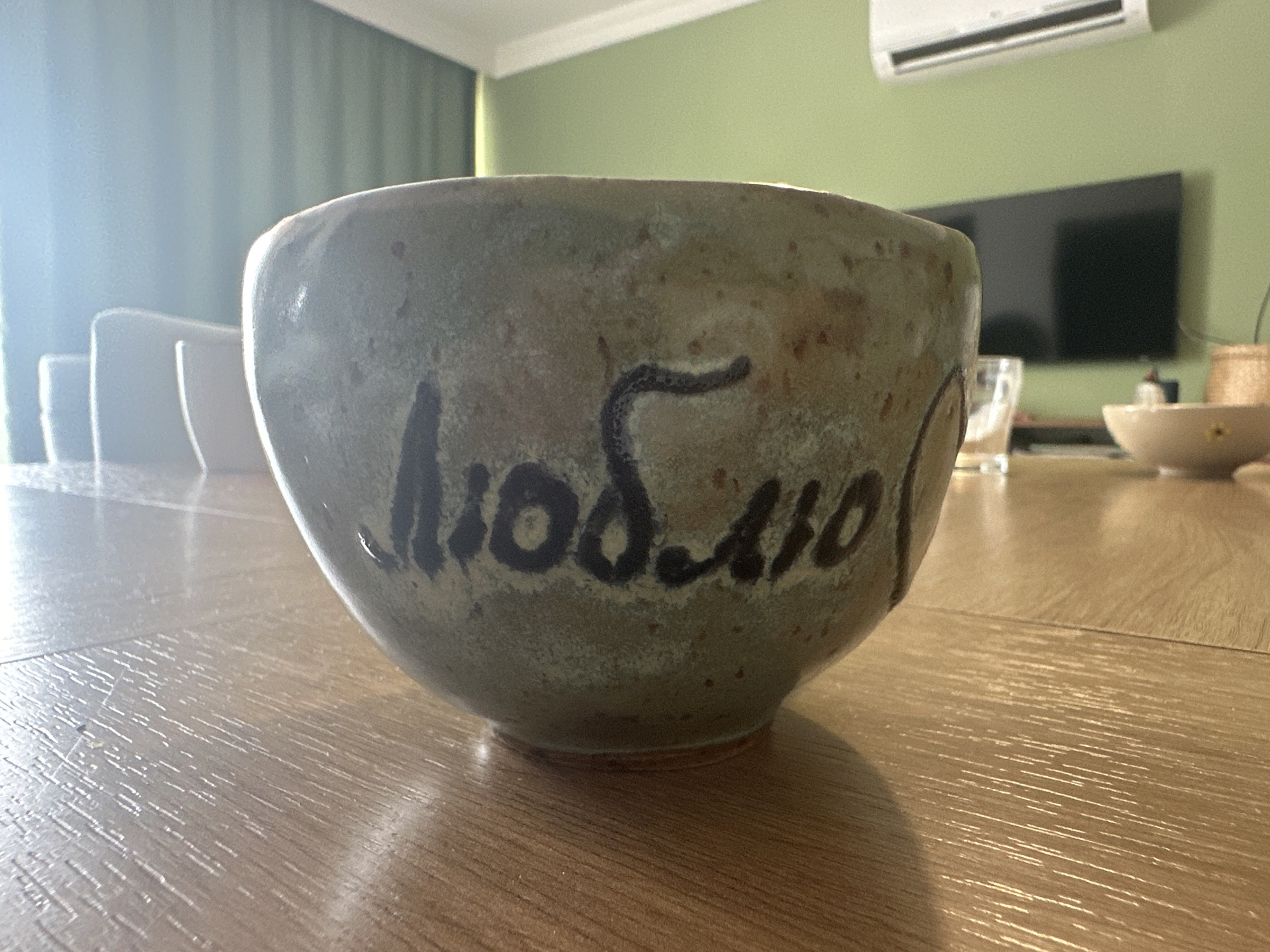 The image depicts a ceramic bowl sitting on a light brown wooden surface, likely a table. The bowl is light greenish-gray with dark brown spots and has the word "Люблю" (Russian for "I love") inscribed in dark brown. In the blurred background, there is a modern living room setting. A white sofa is visible on the left. Further back, there's a green wall, a black television, and an air conditioning unit mounted high on the wall. Other bowls and a drinking glass are also present on the table, adding to the domestic setting.
While the image has a domestic setting, it does not provide specific details about the location, making it impossible to definitively place it in Fethiye, Turkey. The image could be taken anywhere.